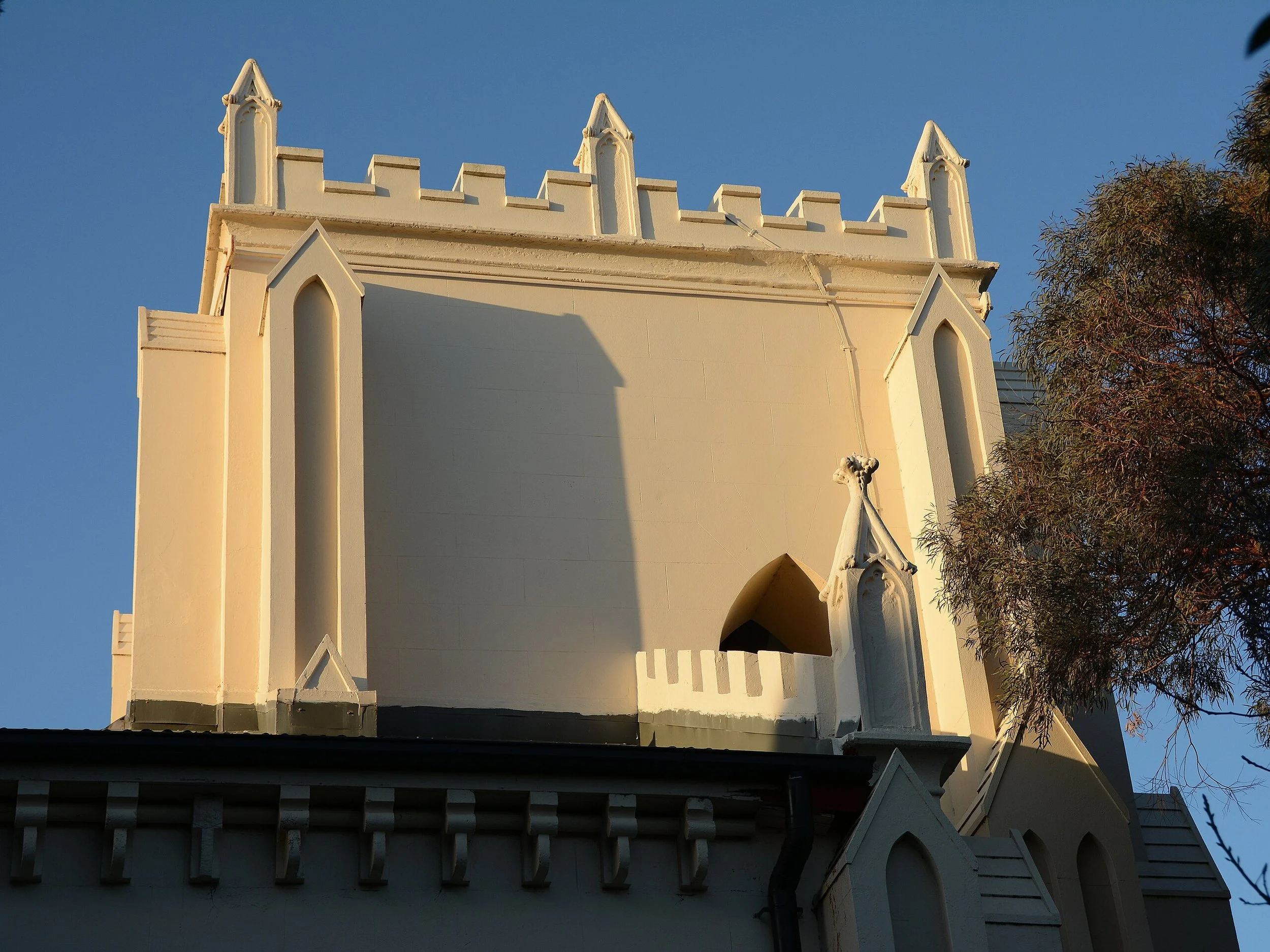 Close-up of a white building with Gothic architectural features, including pointed arches and decorative battlements, under a clear blue sky. Part of a tree with dark green leaves is on the right side.