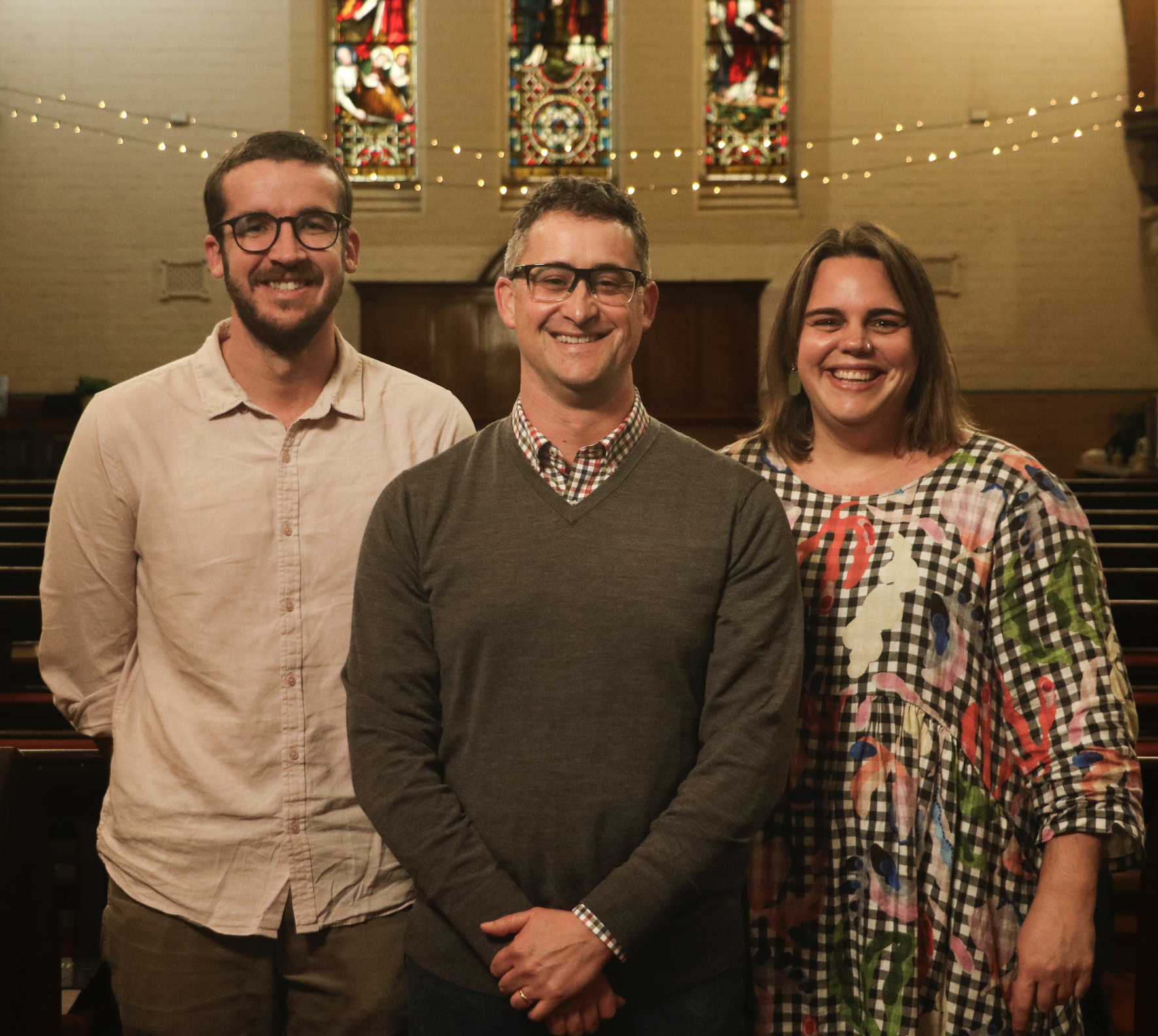 Three people standing inside a church with stained glass windows and string lights, smiling at the camera.