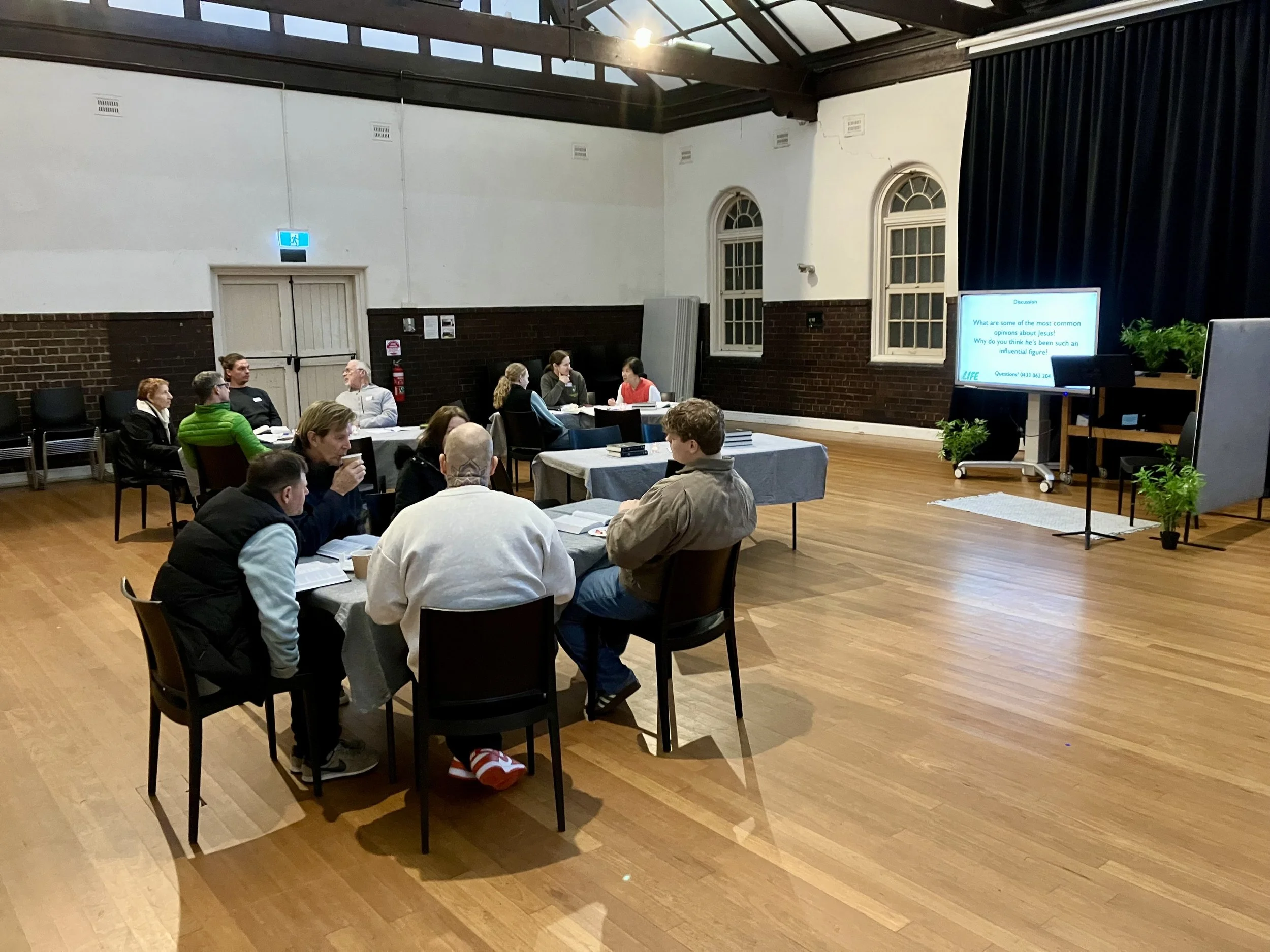 Group of people seated around tables in a room, with a large screen on the right displaying a discussion question. The room has a high ceiling with skylights and brick walls.