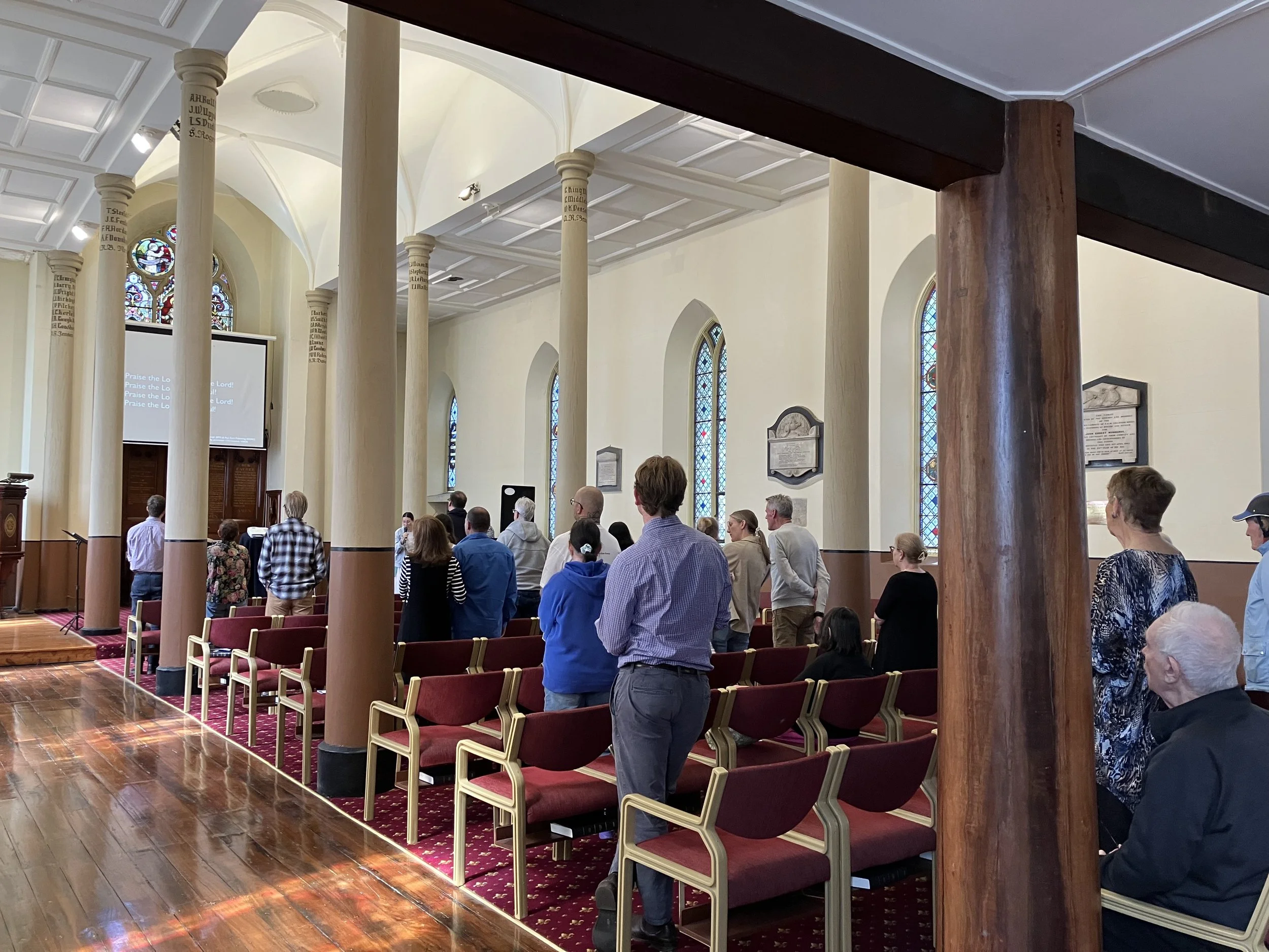 People gathered inside a church during a service, with stained glass windows, wooden beams, and red cushioned chairs.