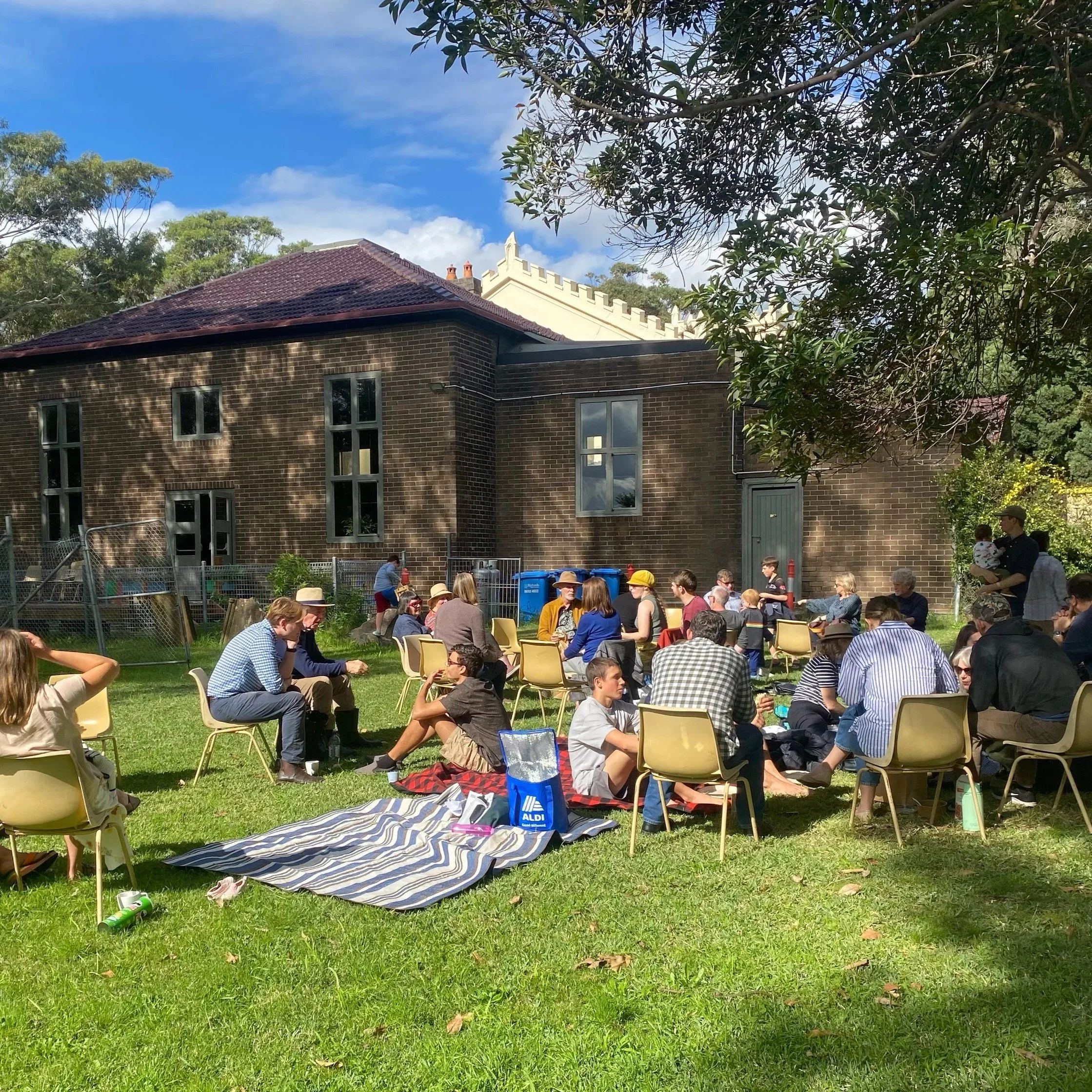 A large gathering of people sitting on yellow chairs outdoors on a grassy area, with some people standing and talking. In the background, there is a brick building with multiple windows, surrounded by trees. The sky is partly cloudy with patches of blue visible.