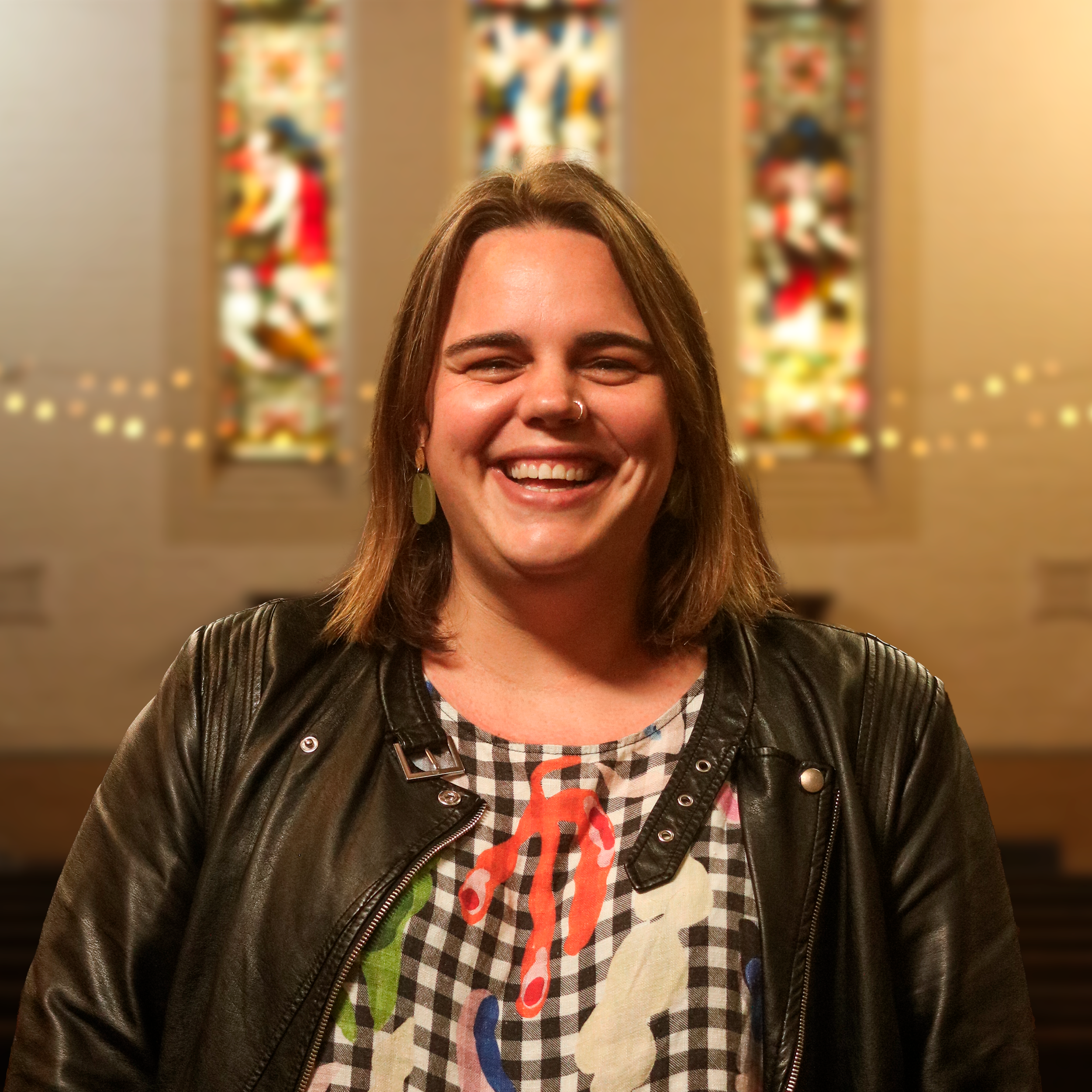 Close-up of a smiling woman with shoulder-length hair, wearing a leather jacket, in front of colorful, abstract artwork.
