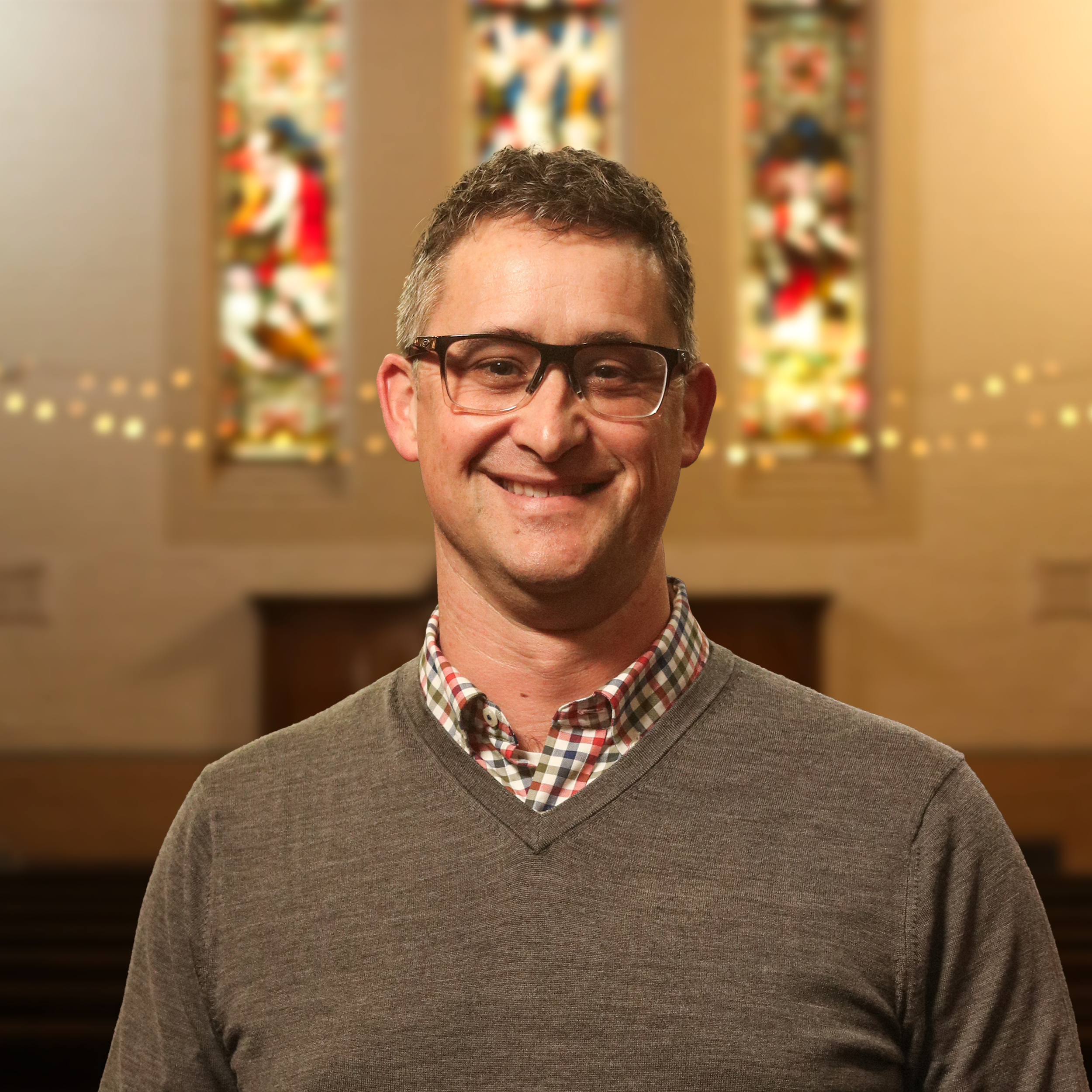 A smiling man with glasses and short, curly hair wearing a brown sweater over a checkered shirt, standing in front of colorful, abstract artwork with blurred lights in the background.