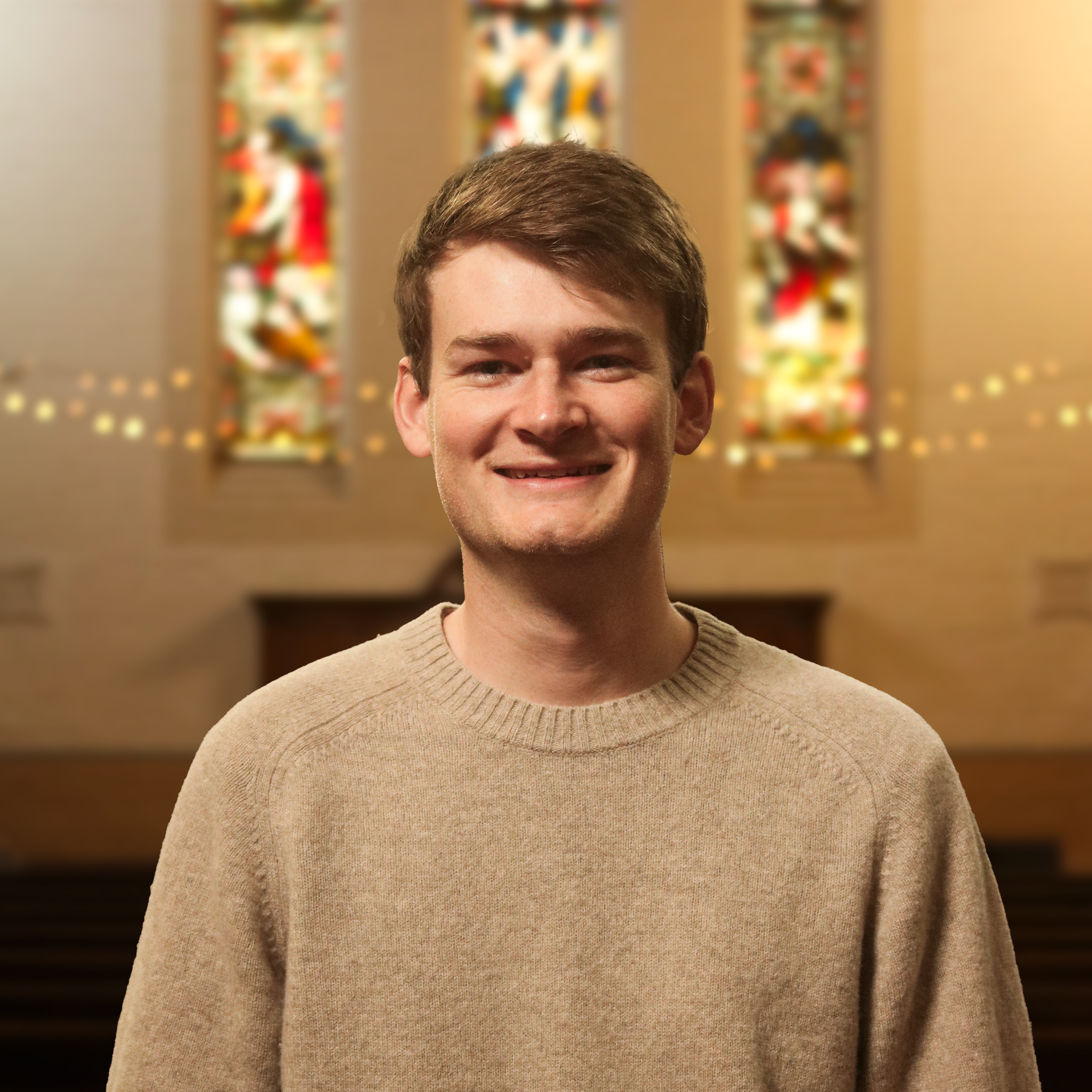 Close-up of a young man with short brown hair smiling, wearing a beige sweater, standing indoors with blurred colorful artwork in the background.