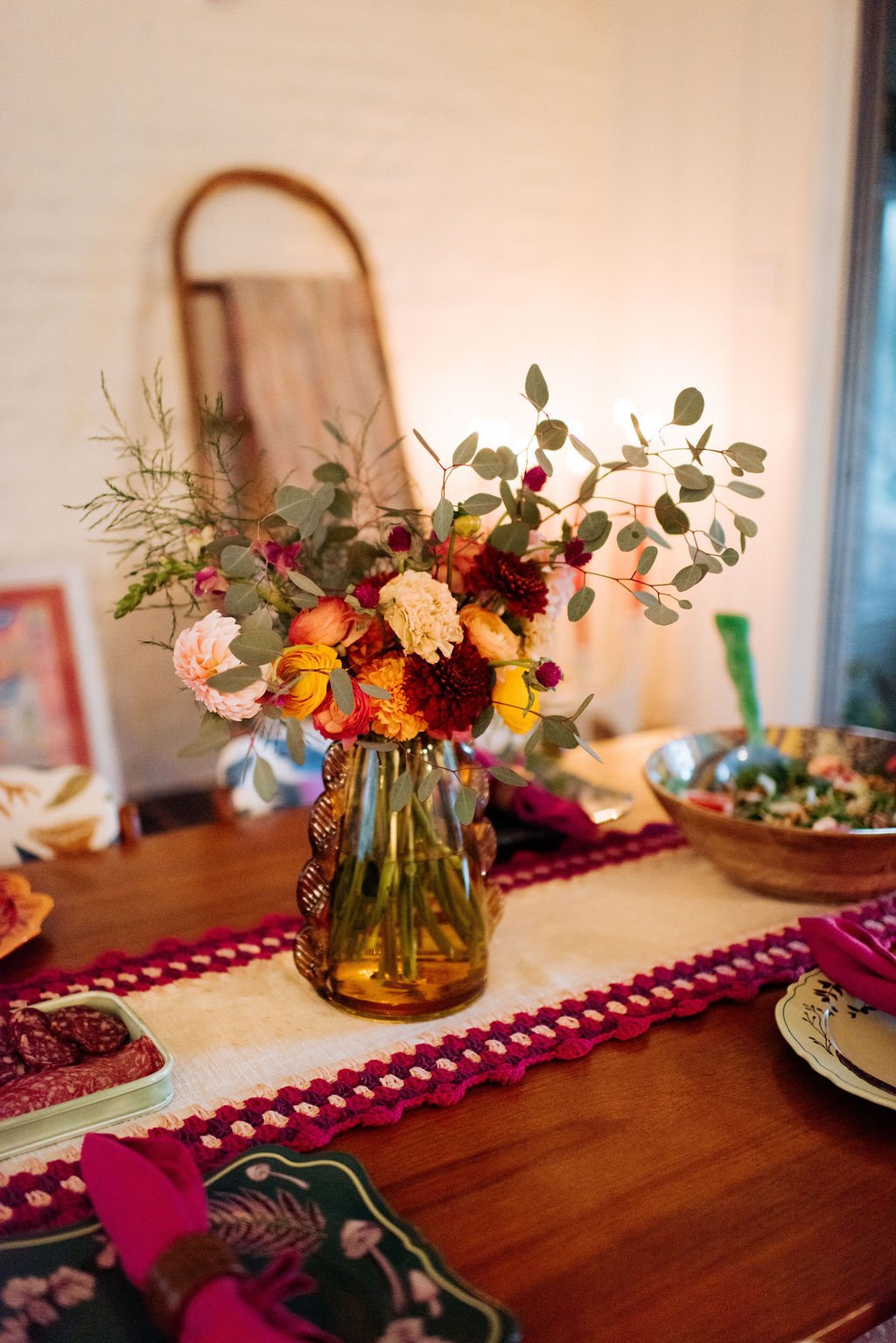 A floral centerpiece with various colorful flowers and greenery in a glass vase on a dining table, with a mirror and a bowl of salad in the background.