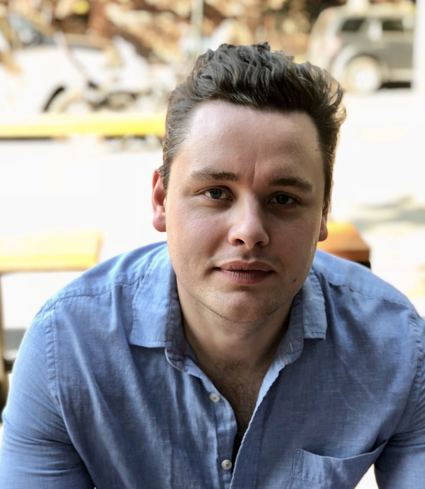 A young man with dark hair and light skin wearing a blue shirt, sitting outdoors, with a blurred background including parked cars and trees.