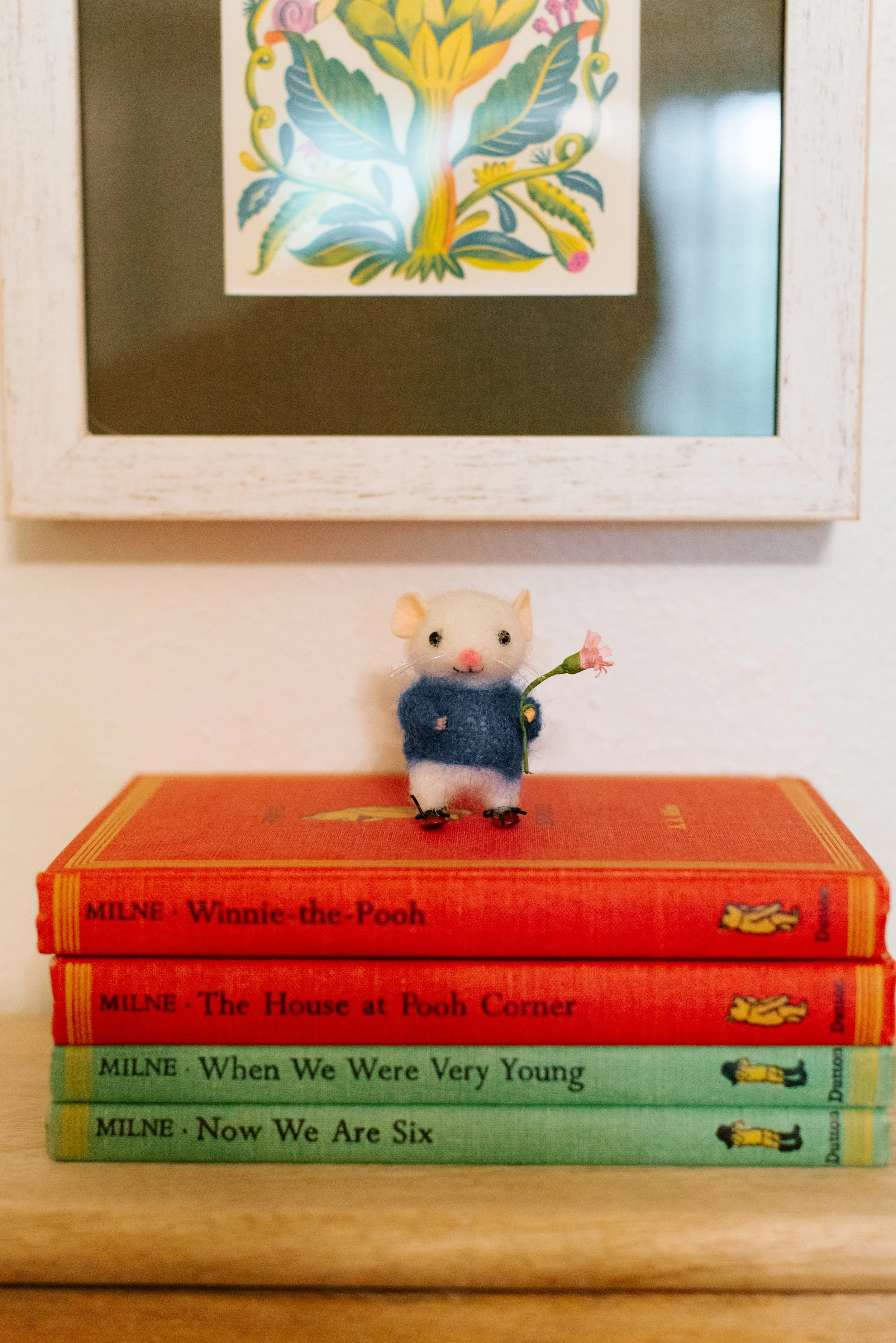 A small plush mouse holding a pink flower stands on a stack of four children's books about Winnie the Pooh, with a framed colorful flower artwork hanging on a wall behind.