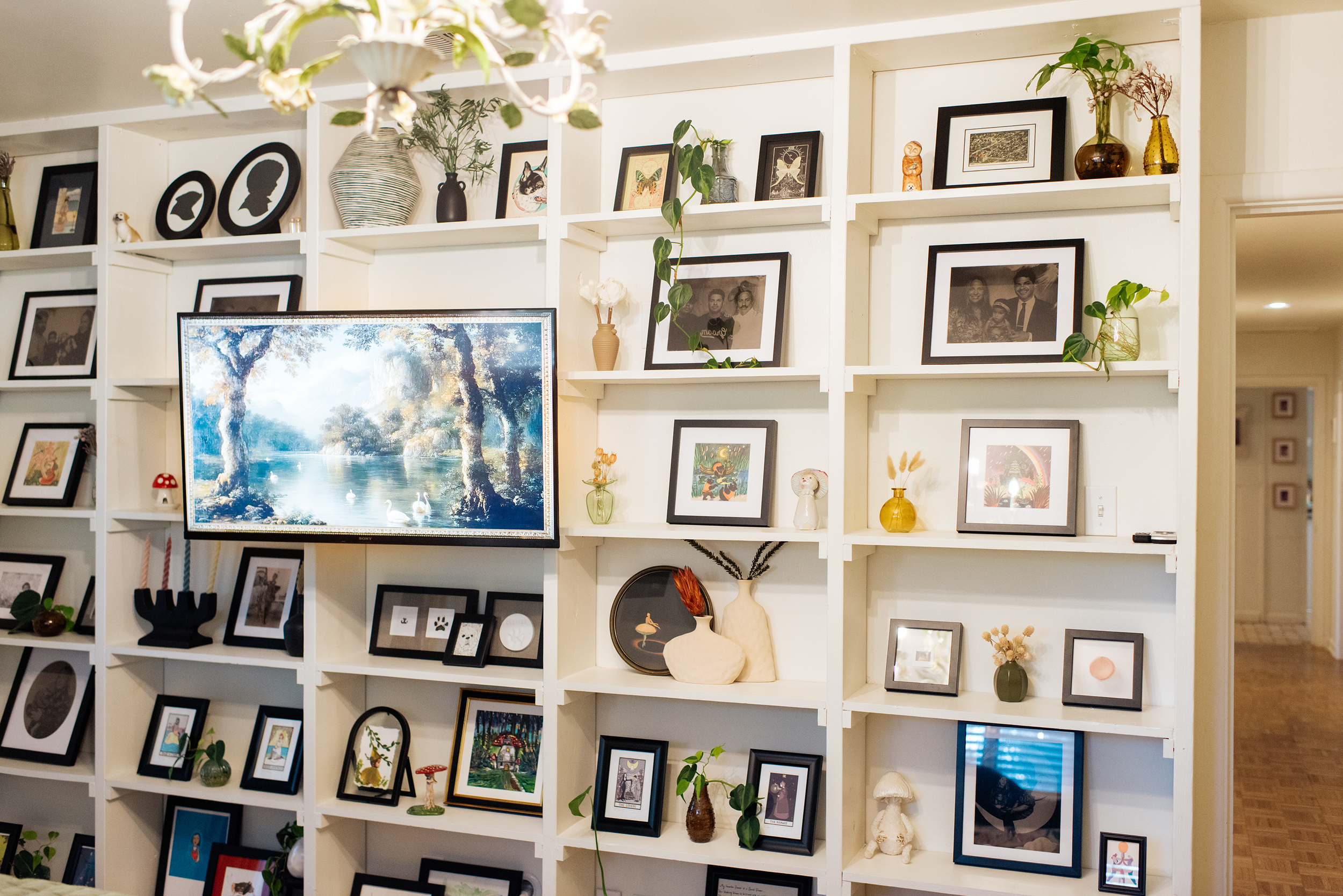 A wall filled with shelves holding framed photographs, artwork, and decorative items, above a mounted television displaying a nature scene with trees and swans.