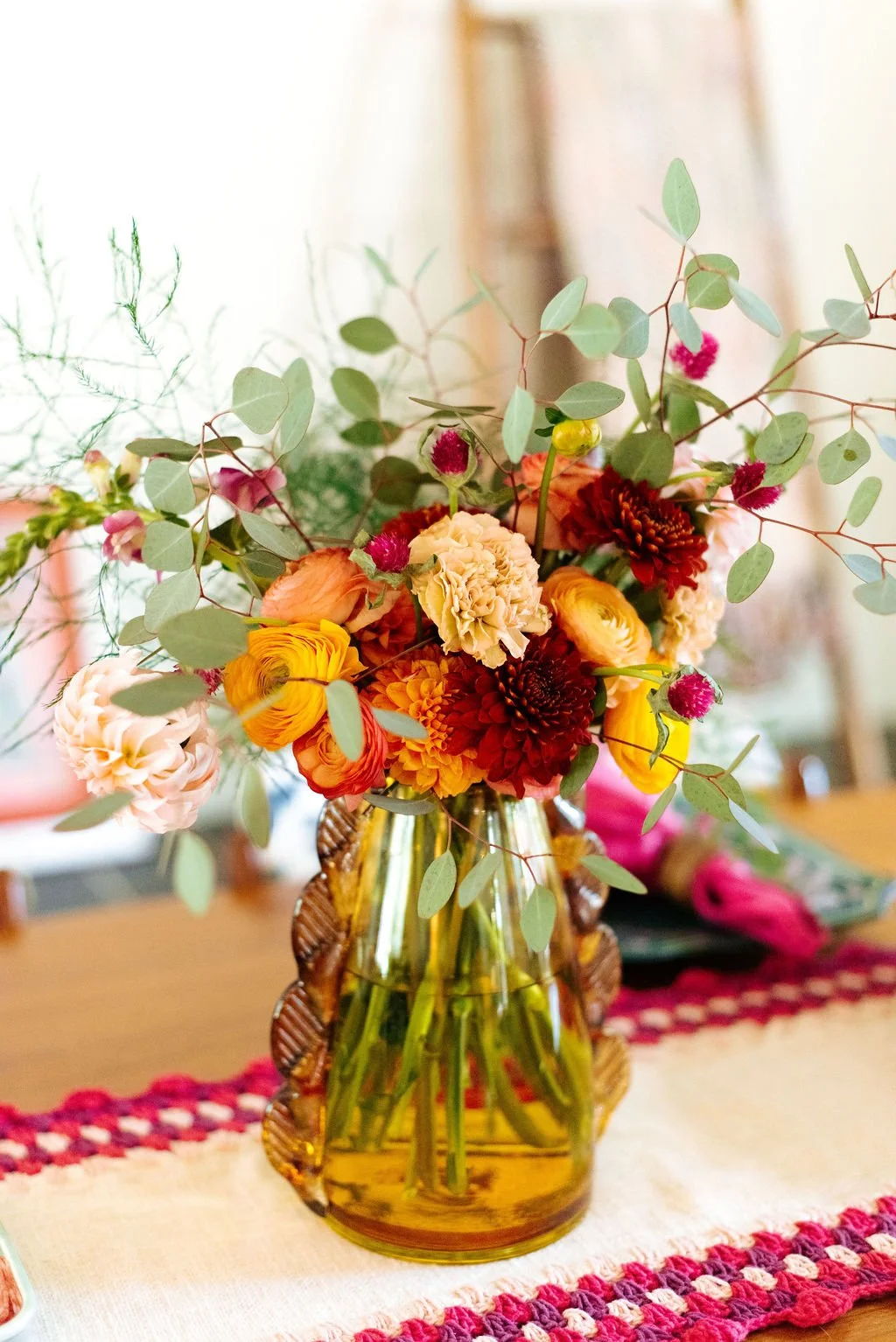 A colorful bouquet of flowers in a yellow glass vase placed on a table with a red and white crocheted runner.