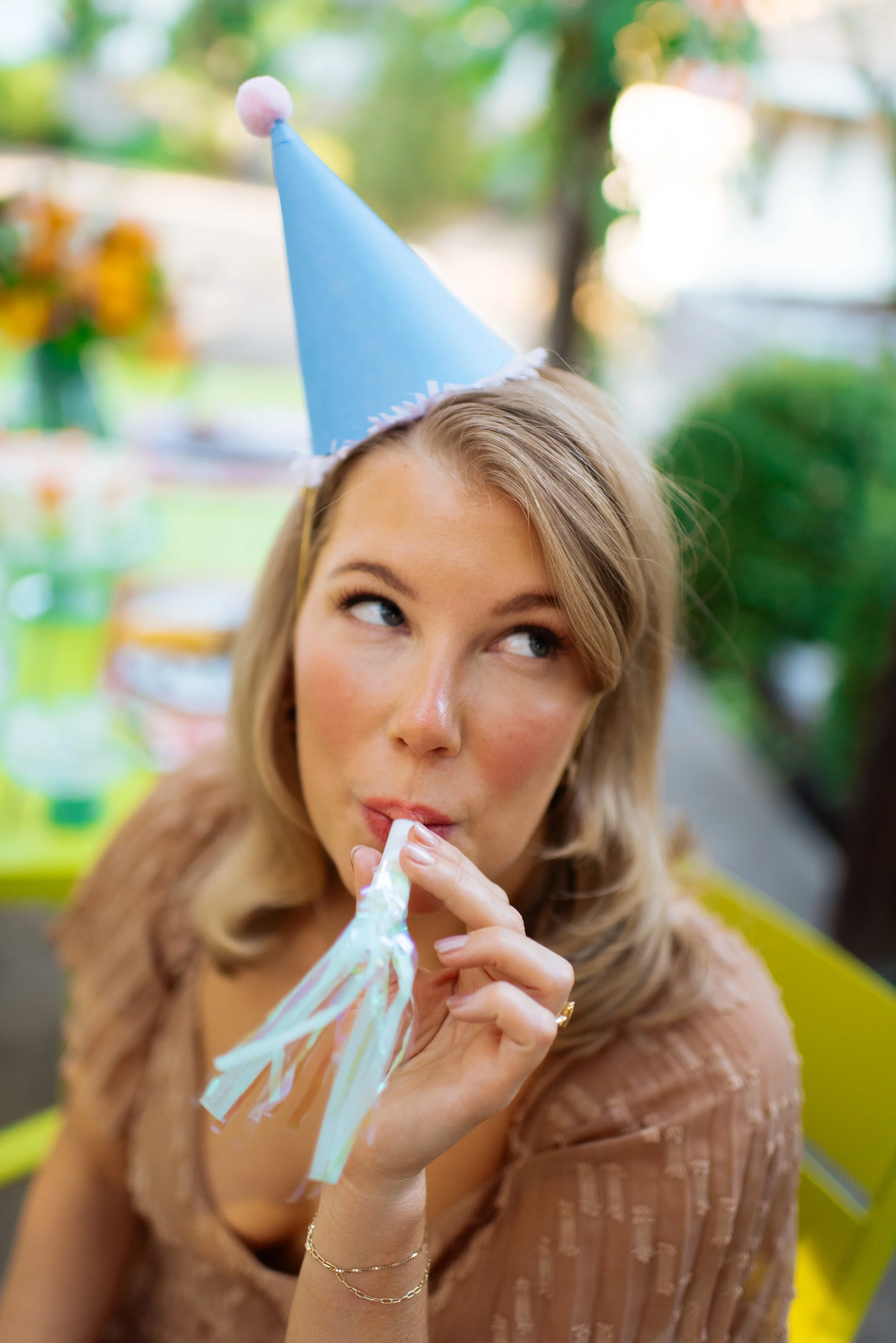 Young woman wearing a blue birthday cone hat with pink pom-pom, holding a party streamer near her lips, with blurred colorful party decorations in the background.