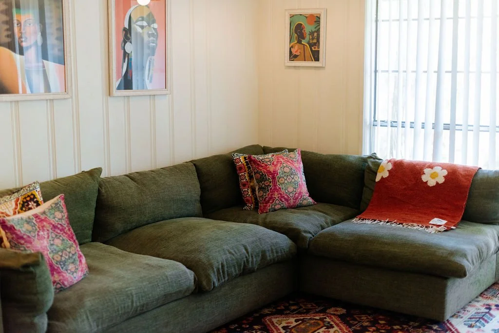Living room with a dark green sectional sofa, decorative pillows, colorful artwork on the white paneled walls, and a window with white curtains.