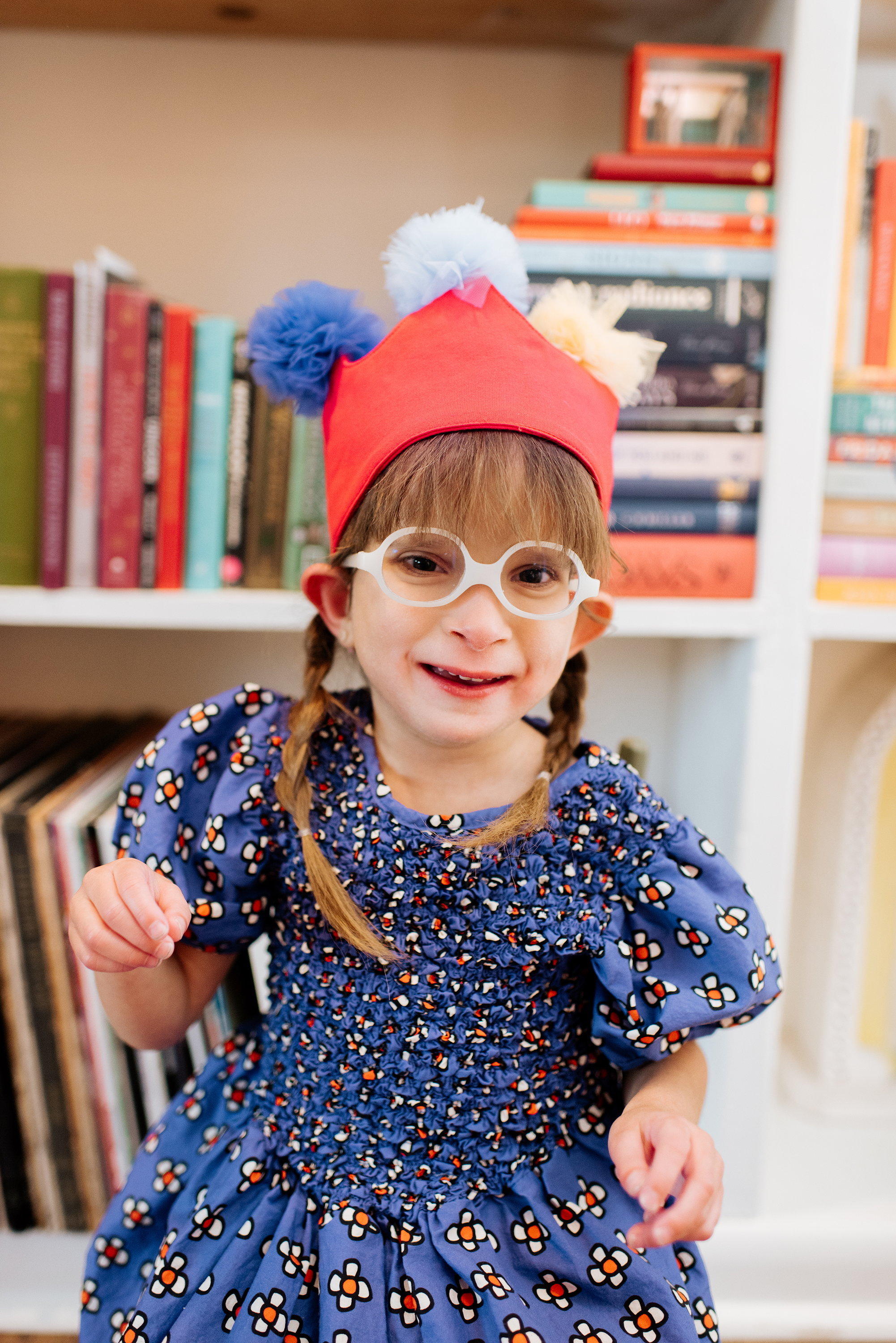 A young girl wearing a red clown hat with pom-poms, glasses, and a blue dress with a floral pattern, smiling and standing in front of a bookshelf.
