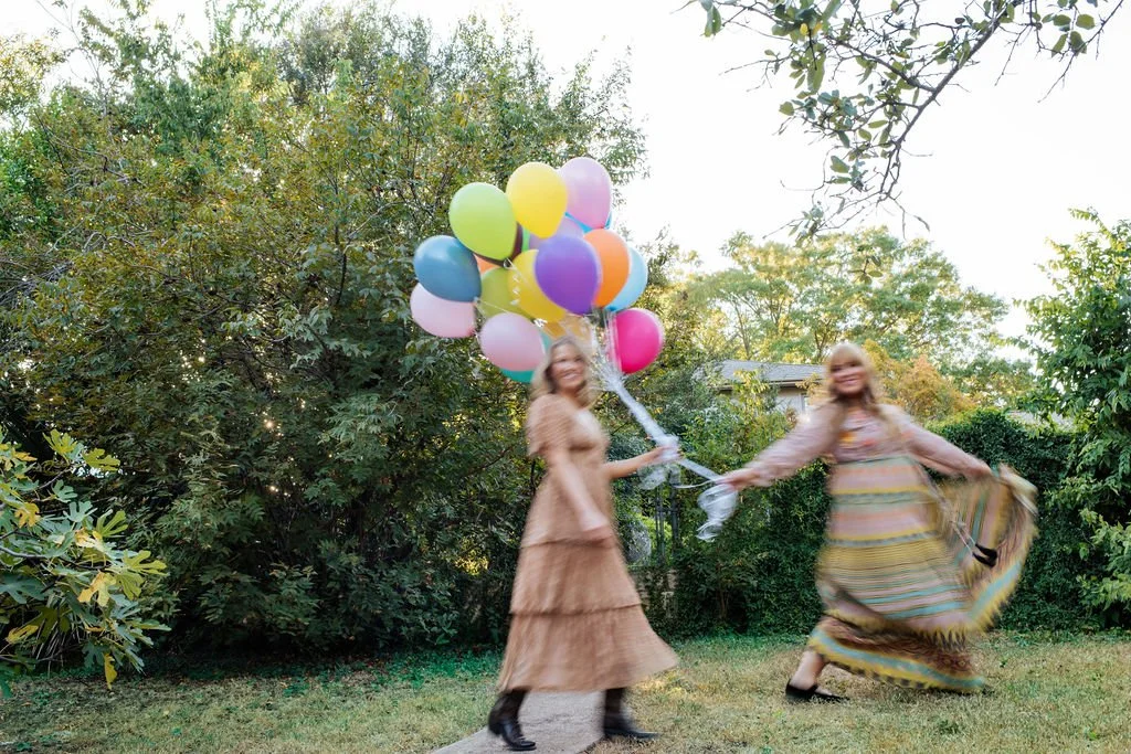 Two women in vintage-style dresses holding a bunch of colorful balloons in a garden with trees.