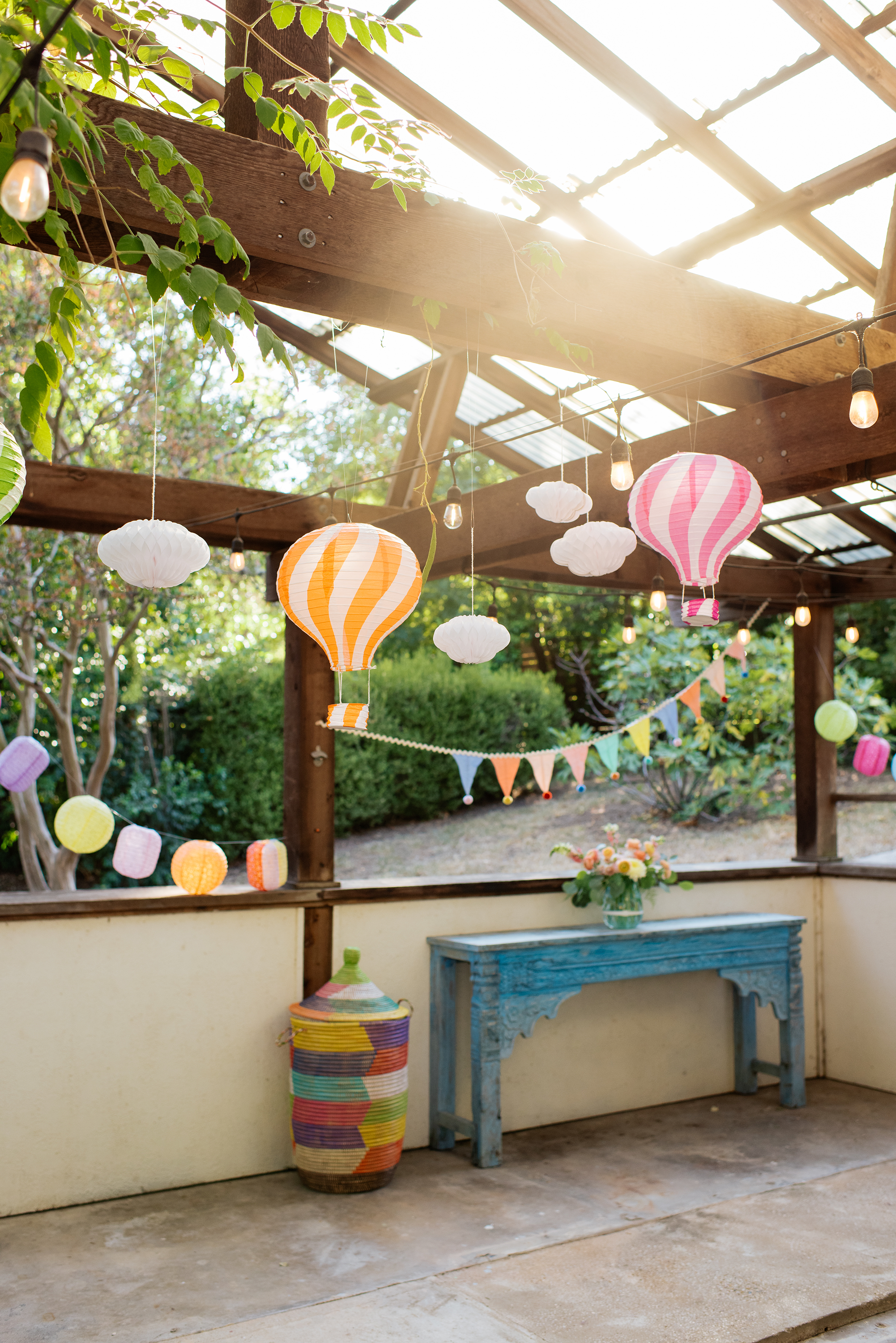 Decorative outdoor patio or porch area with hanging string lights, paper lanterns, and bunting flags, with a blue distressed wood table holding a flower arrangement.