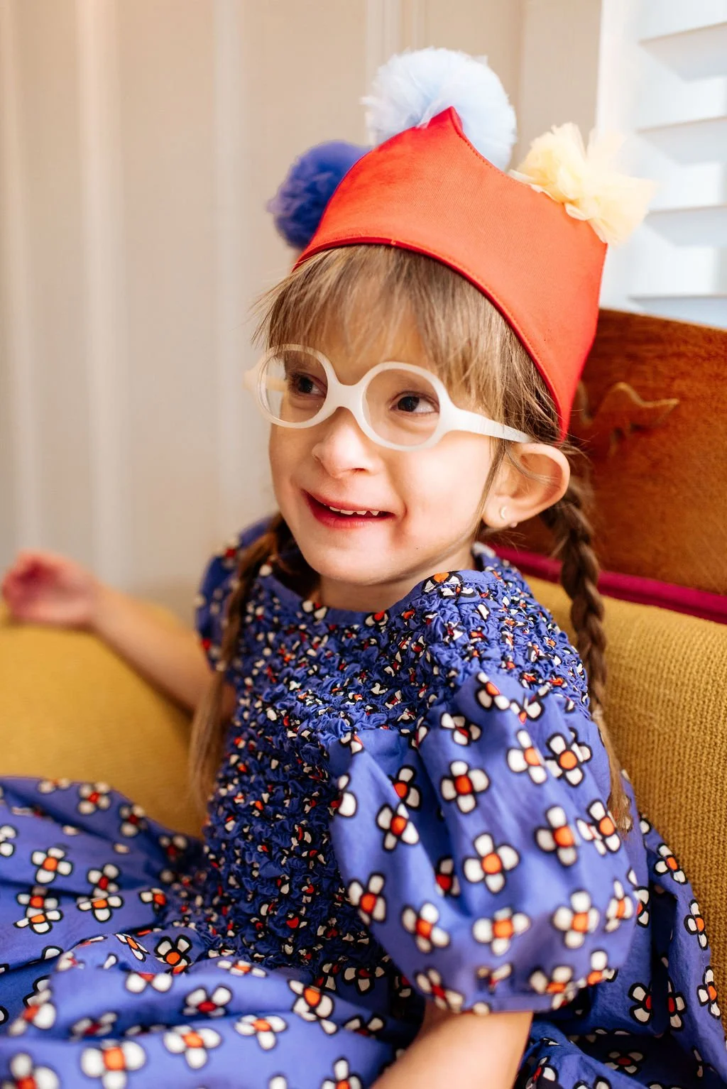 A young girl with braided hair wearing white glasses and a colorful clown hat, smiling and sitting on a yellow couch in a cozy indoor setting.