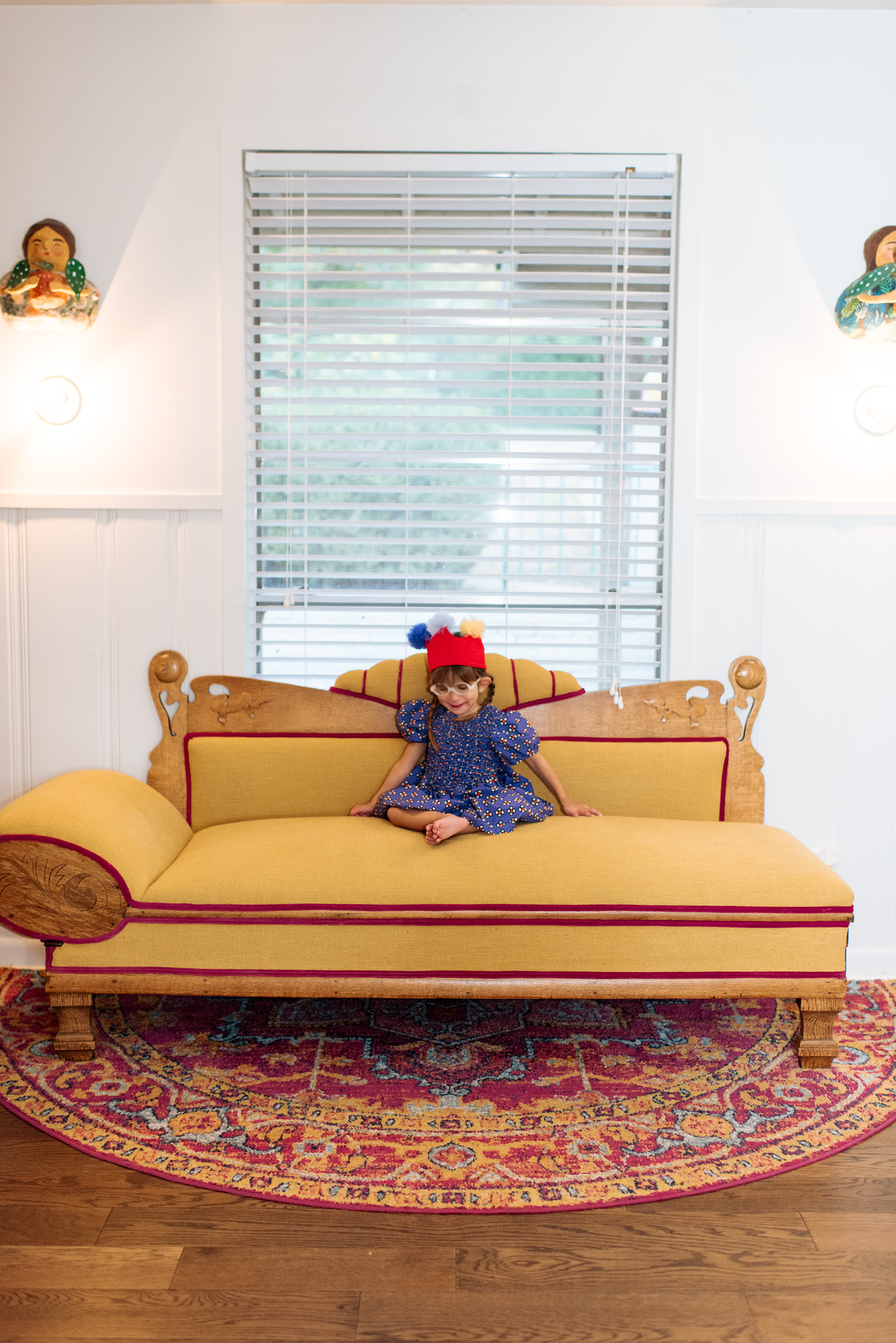 A young girl sitting on a yellow vintage-style couch with wooden accents and red trim, wearing a blue dress with colorful patterns and a red hat with pom-poms, in front of a window with blinds in a well-lit room.