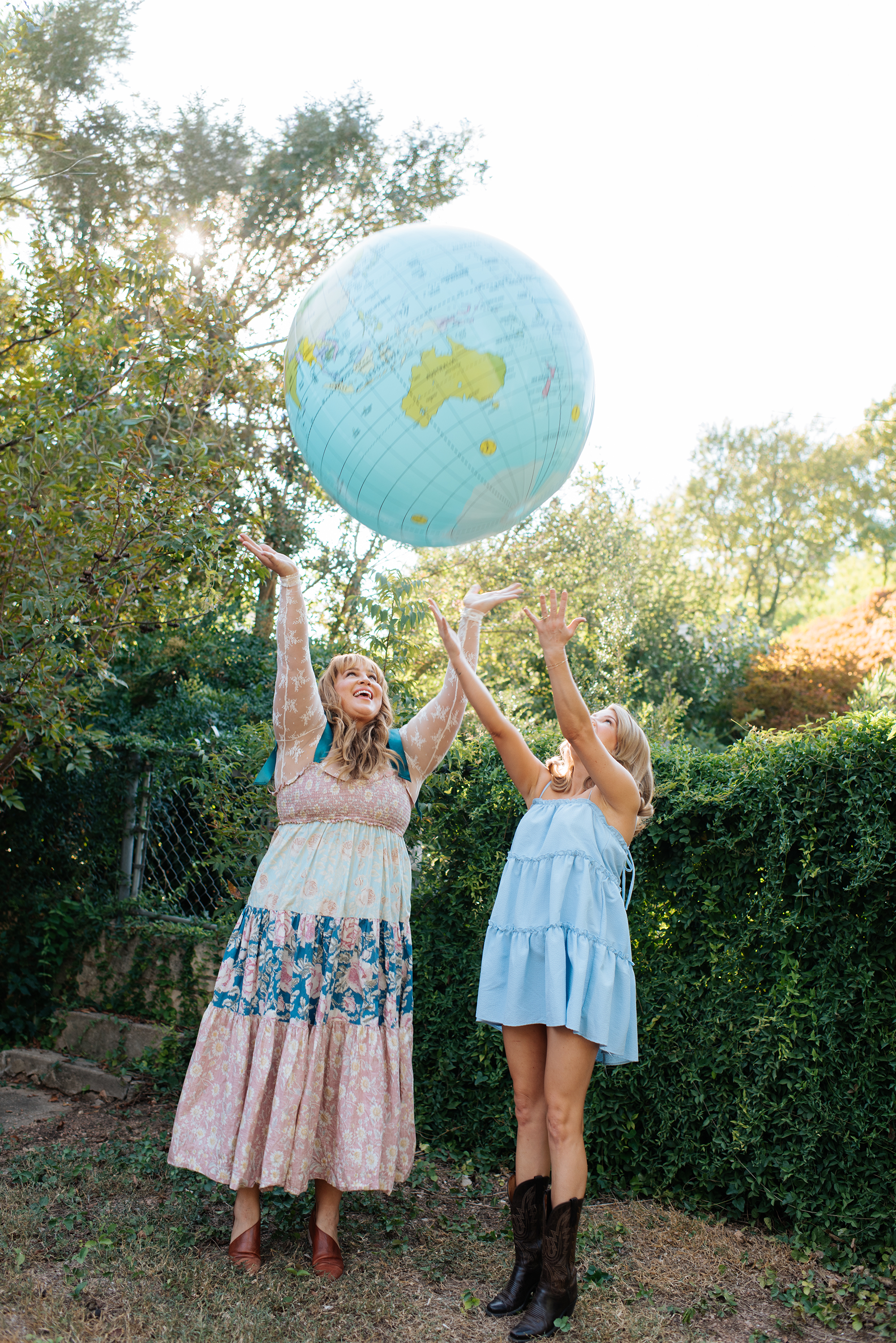 Two women in summer dresses playing with a large inflatable globe outdoors among trees on a sunny day.