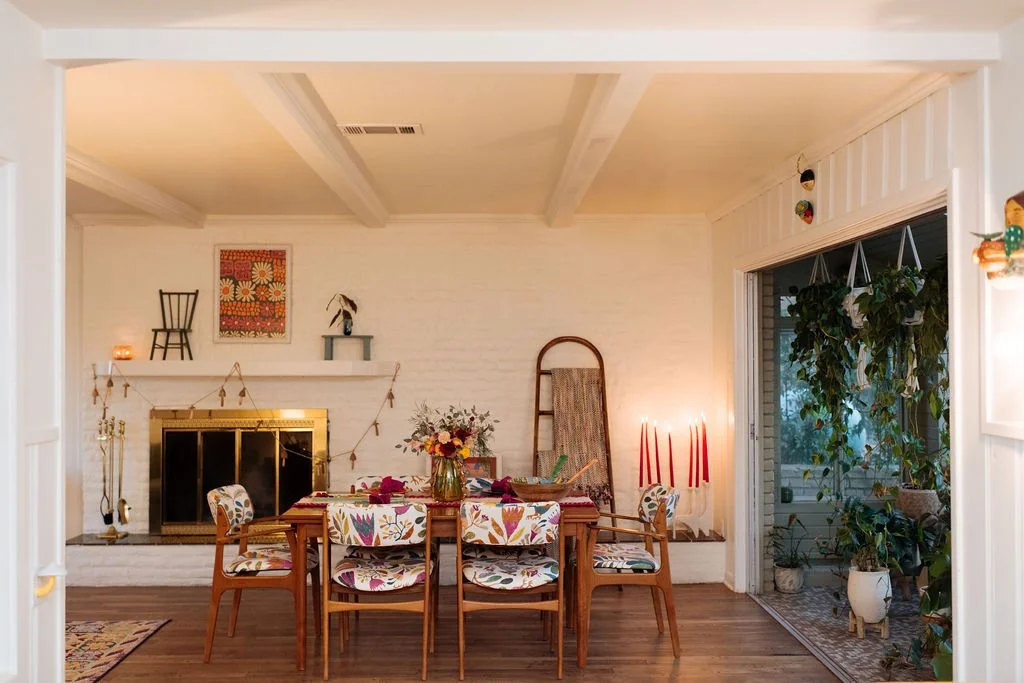 A cozy dining room with a wooden table, floral upholstered chairs, and a decorated fireplace with a string of lights. There's a tall scroll back chair, a flower vase, and red candles beside a glass sliding door leading to a lush green porch.