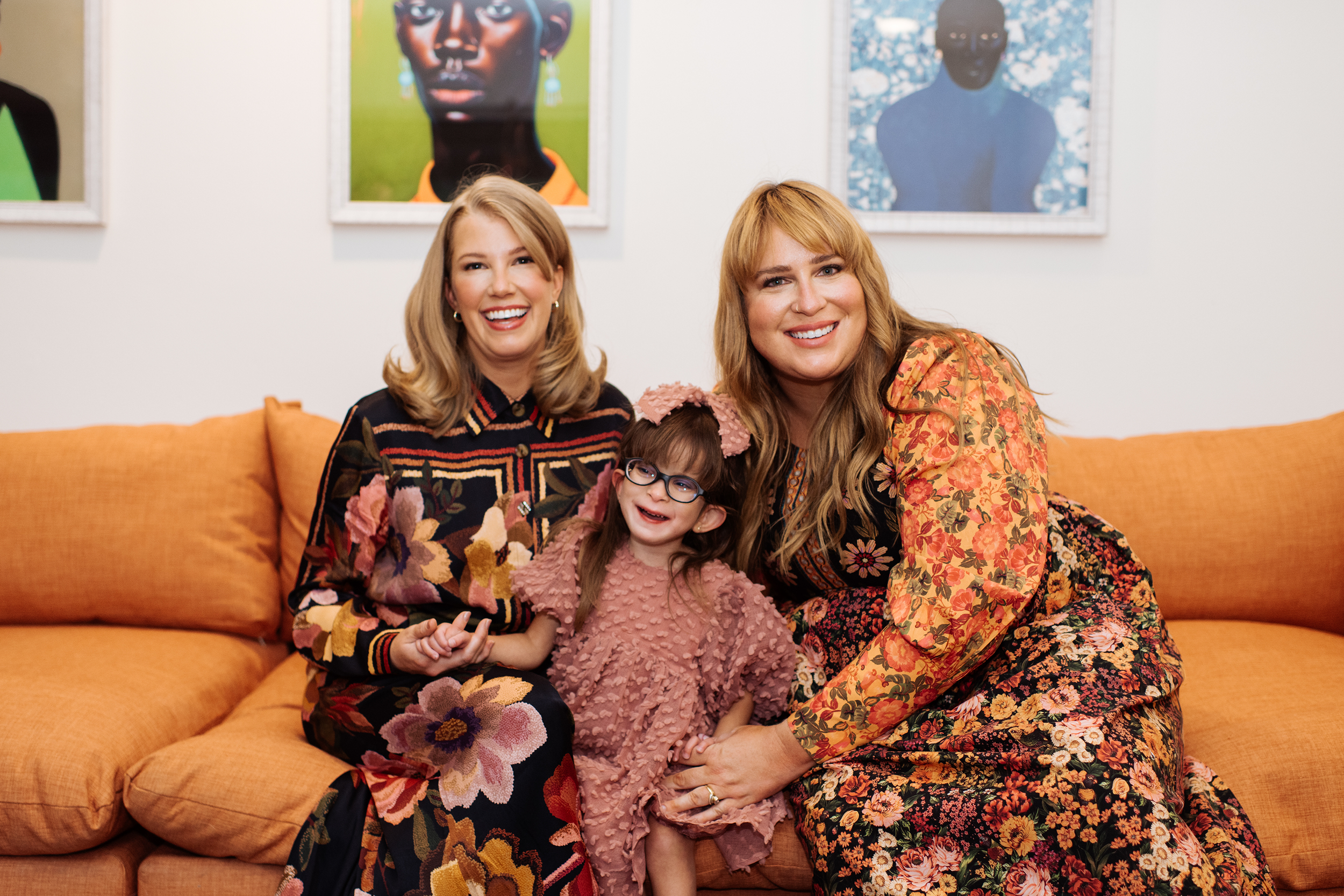 Two women and a young girl sitting on an orange couch, smiling at the camera, with colorful artwork on the wall behind them.