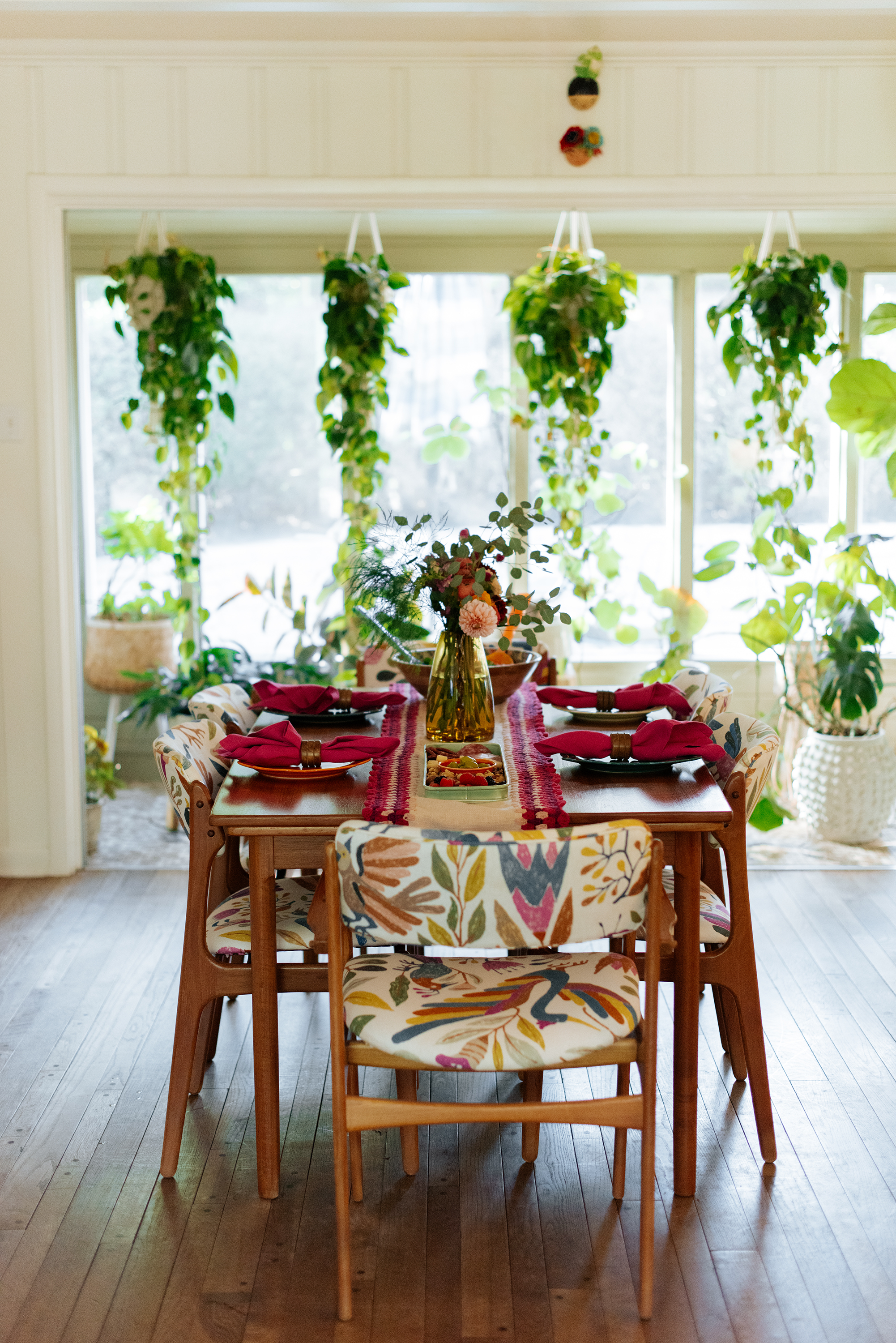 A cozy dining area with a wooden table set for a meal, featuring red napkins, a floral centerpiece in a yellow vase, and surrounded by six upholstered chairs with colorful, nature-inspired patterns. The background shows a large window with hanging pl