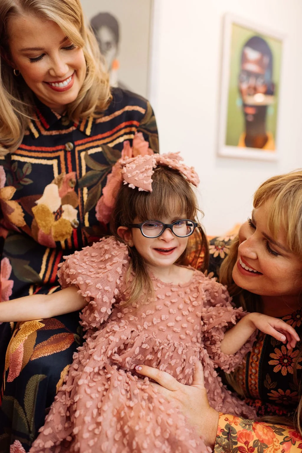 A young girl with glasses and a pink textured dress is smiling, with two women and another person partially visible around her. One woman is smiling at her, and the other woman is smiling while holding her. The girls and women are in a room decorated