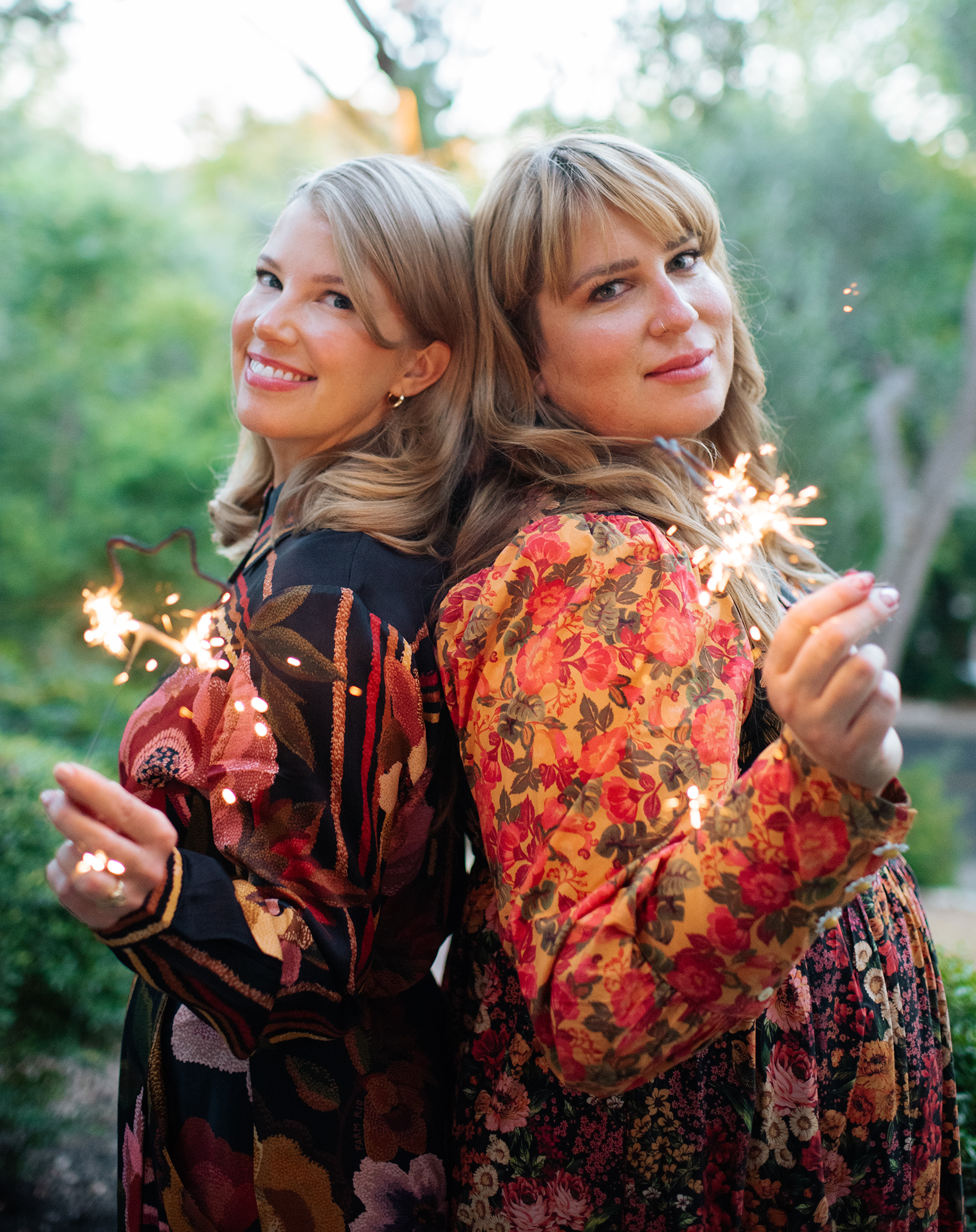 Two women standing back-to-back outdoors, each holding sparklers, smiling, surrounded by trees.