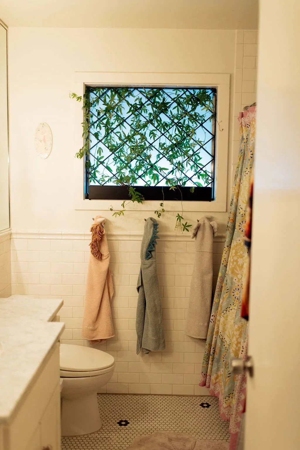 Small bathroom with a window covered by a black lattice and greenery outside. Three towels hang on hooks, and a shower curtain with a colorful floral pattern is partially visible. The bathroom has white tiled walls and hexagonal floor tiles.