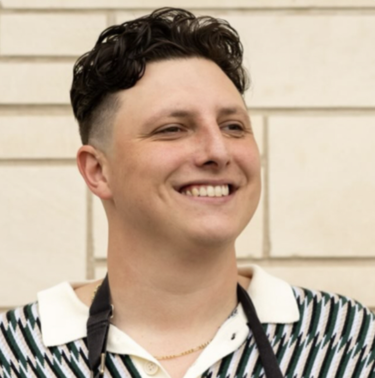 A young man with dark, wavy hair smiling, wearing a striped shirt and a camera strap around his neck, standing in front of a beige brick wall.