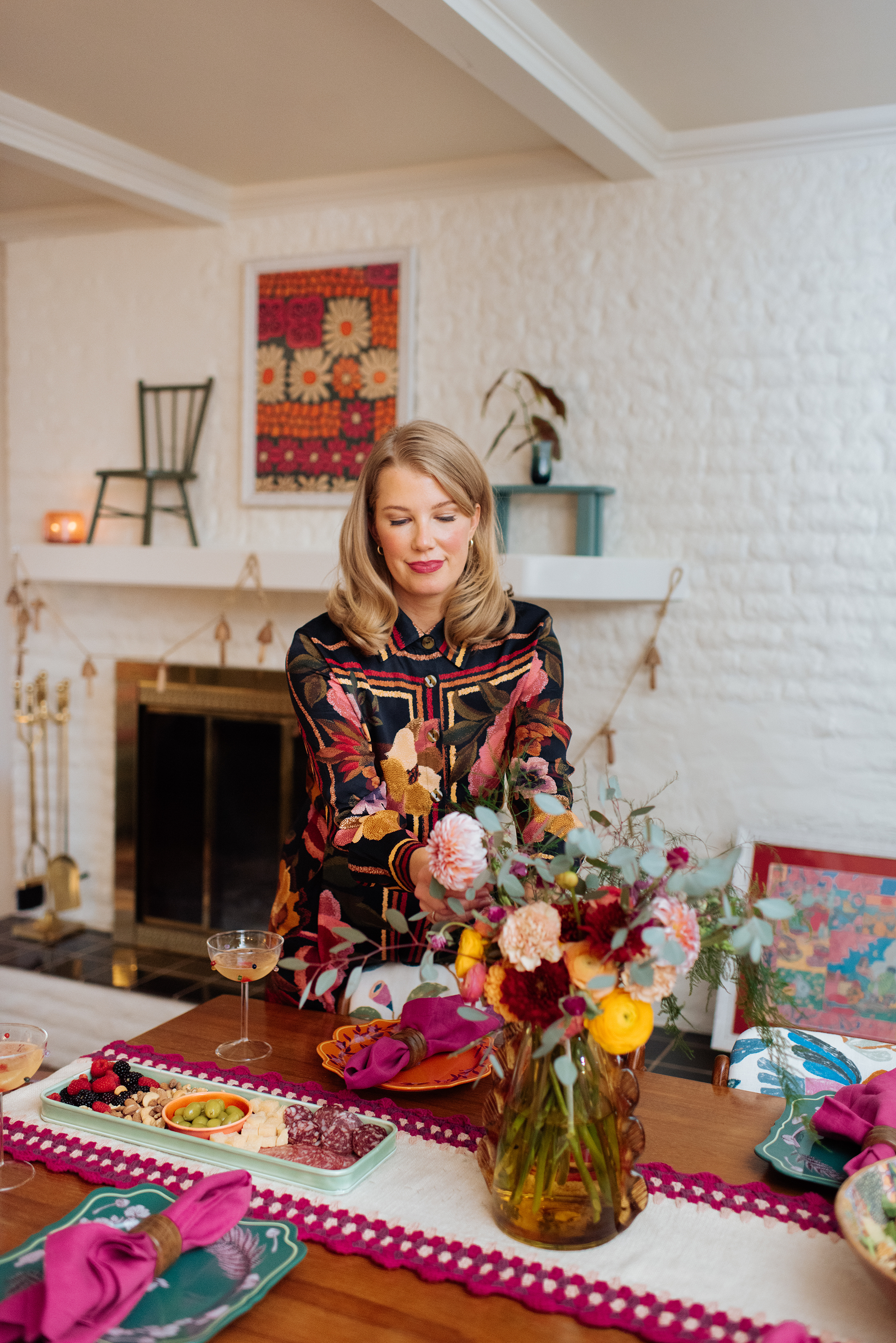 A woman with blonde hair, wearing a floral dress, standing at a decorated dining table with a large flower arrangement and servings of cheese and fruit. The background features a white brick fireplace, a colorful floral artwork, a small chair, and a candle.