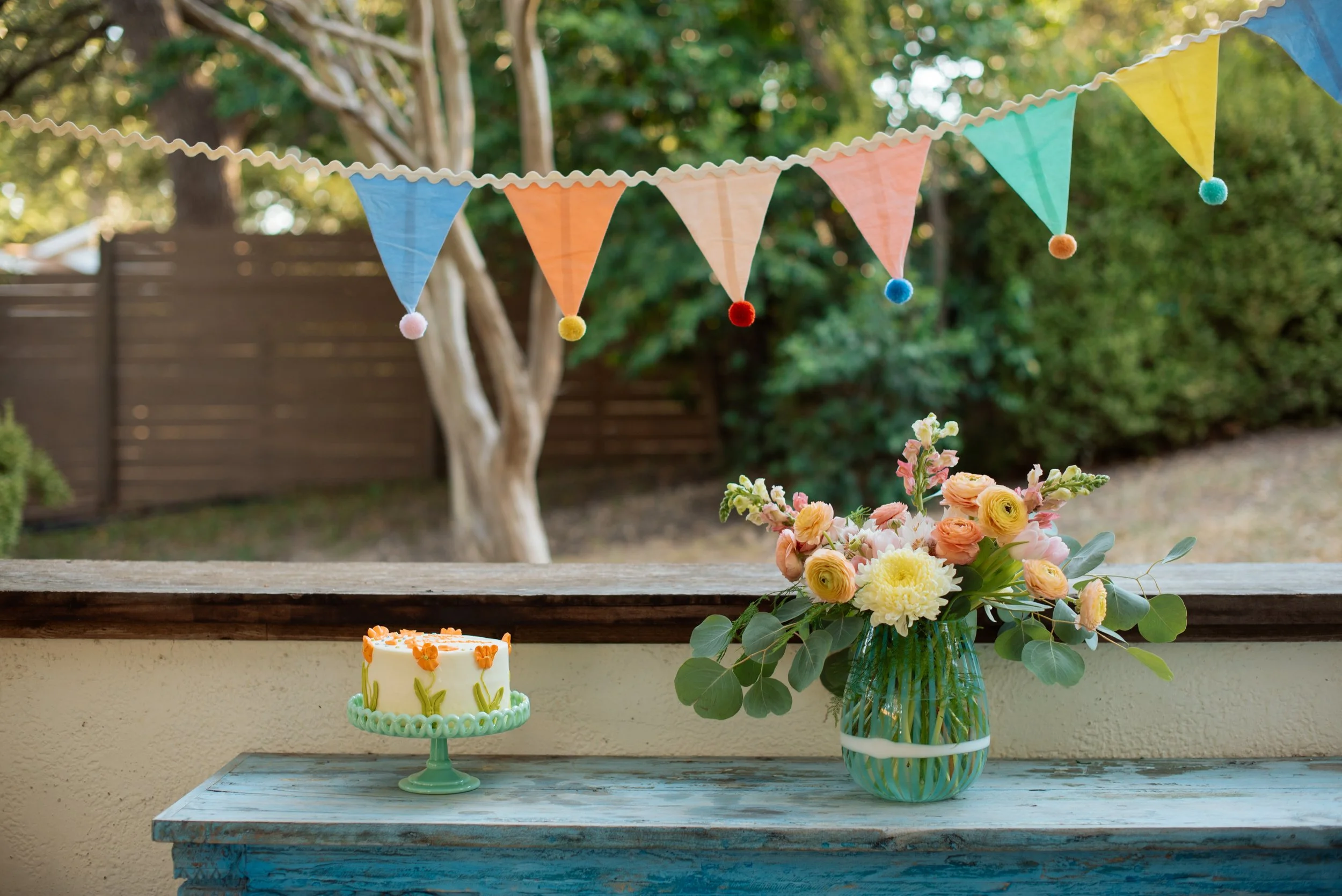 A small decorated birthday cake and a vase of flowers on a blue wooden table, with colorful triangular banners hanging overhead, in an outdoor setting with trees and a wooden fence in the background.