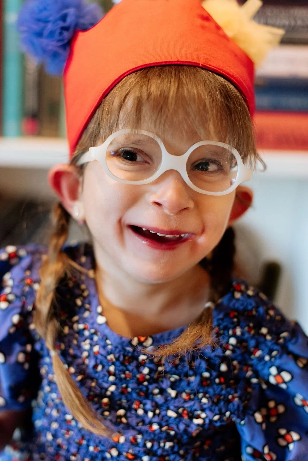 A young girl smiling, wearing white glasses, a red festive hat with pom-poms, and a blue dress with colorful patterns.