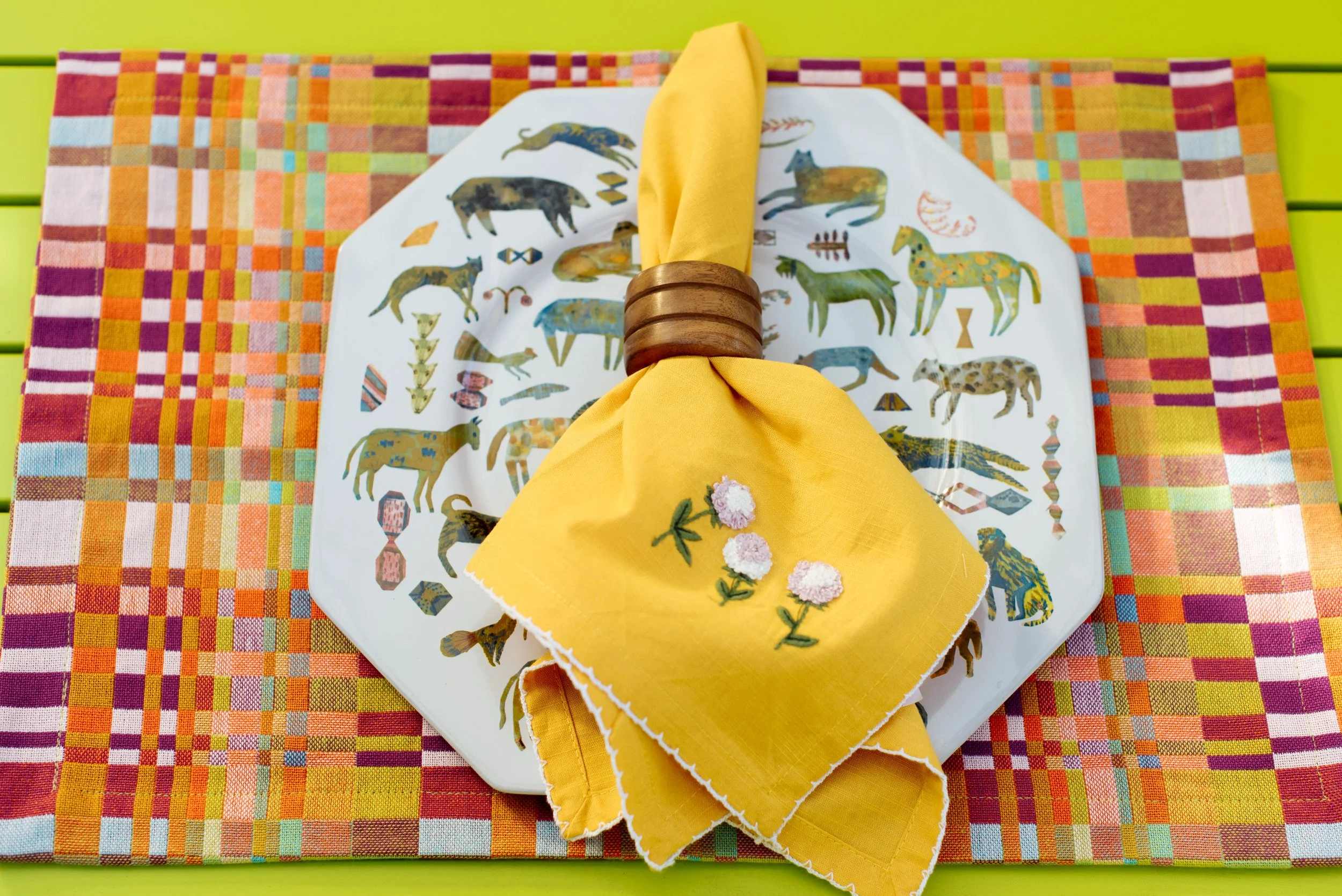 Table setting with a decorative plate featuring images of animals, a yellow napkin with embroidered pink flowers held by a wooden napkin ring, and a colorful woven placemat on a green surface.