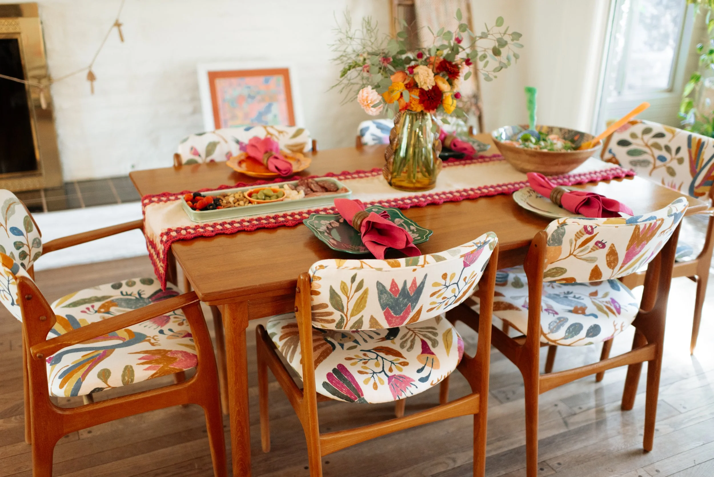 Dining table set with colorful floral chair cushions, a bouquet of flowers in a glass jar, plates with pink napkins, and a runner with pom pom trim, in a bright room with a sliding glass door.