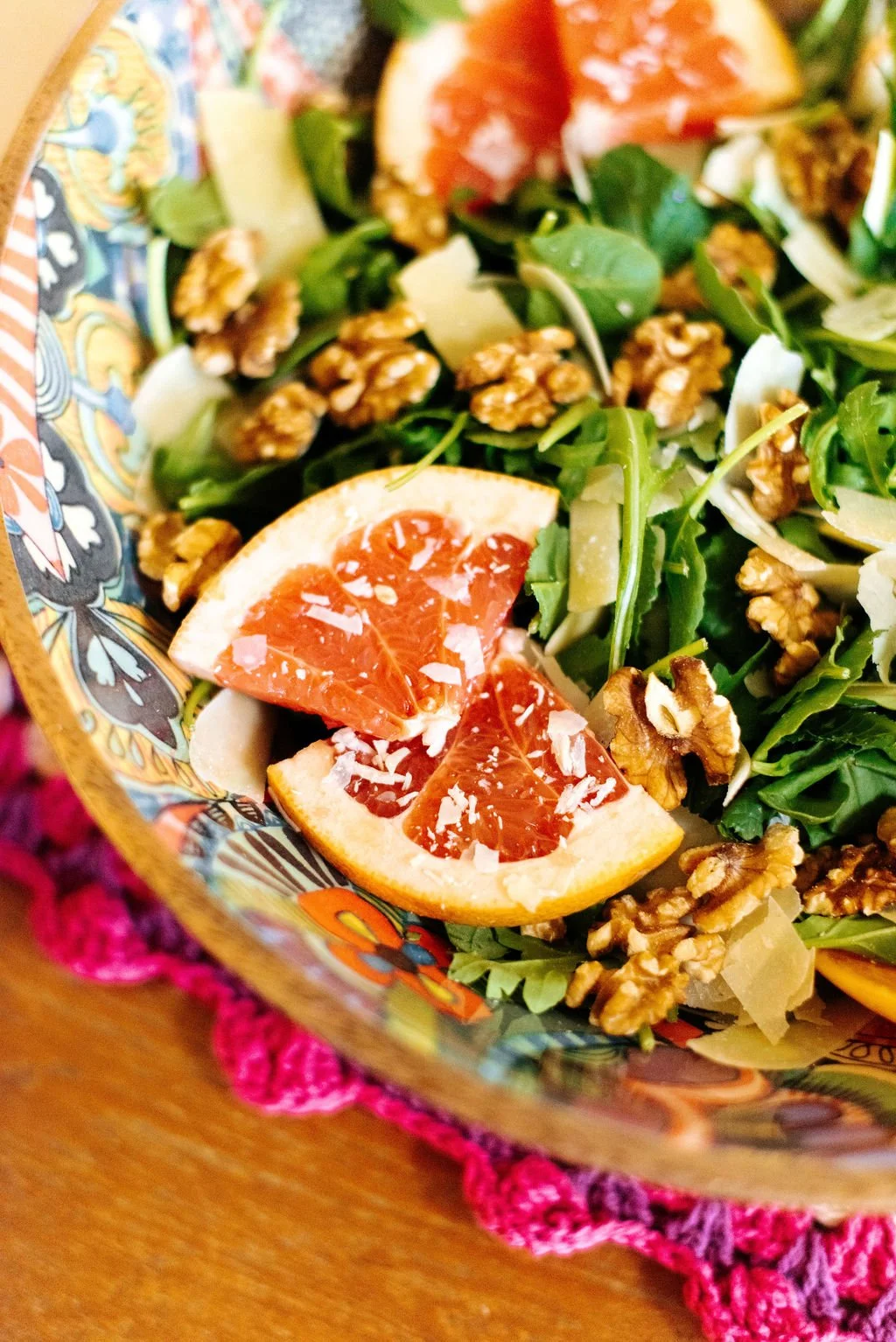 A colorful salad with grapefruit slices, walnuts, greens, and shaved cheese in a decorative bowl on a pink crochet placemat.