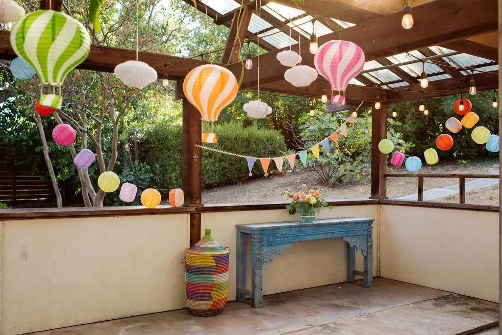 Decorated patio with colorful paper lanterns, string lights, a blue carved wooden table with a flower arrangement, and a woven basket, set in a garden with trees and bushes.