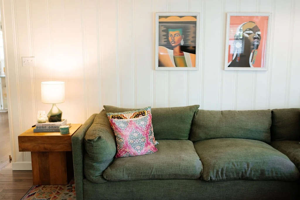 A living room with a green sofa, colorful pillows, a wooden side table with a lamp, a small plant, and some books. Two framed abstract portraits hang on a white paneled wall above the sofa.