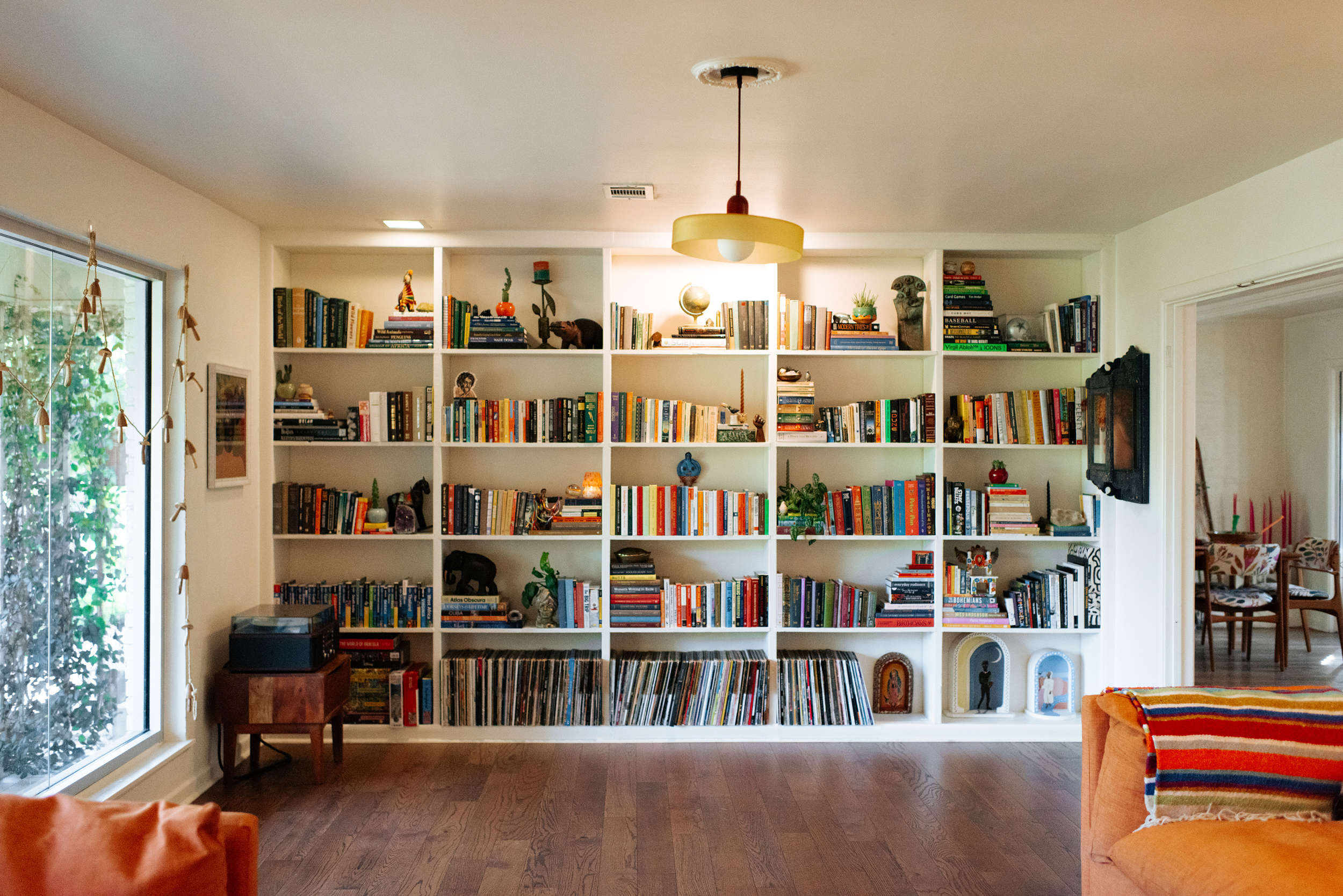 A large white bookshelf filled with books, decorative items, and plants in a living room, with a sliding glass door on the left and a doorway on the right leading to a dining area.