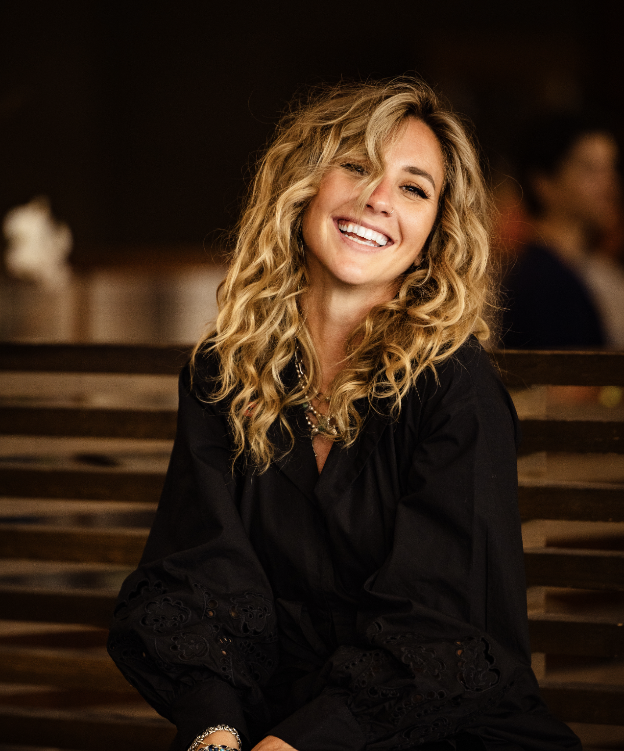 A woman with long, curly blonde hair smiling and looking at the camera, sitting on a wooden bench in a dimly lit indoor setting.