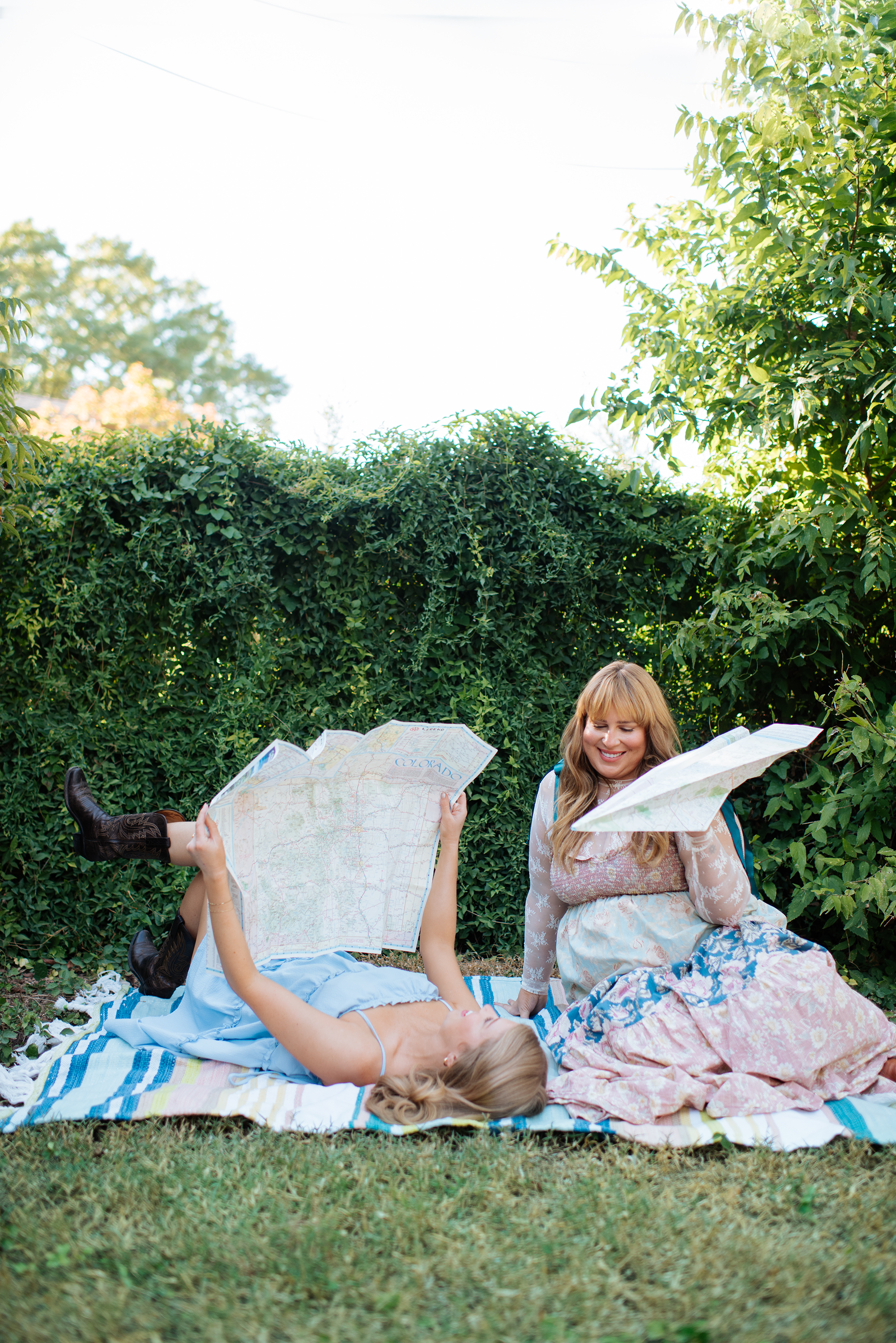 Two women are outdoors on a blanket, one lying down and holding a map, the other sitting and looking at a map, surrounded by green bushes and trees.