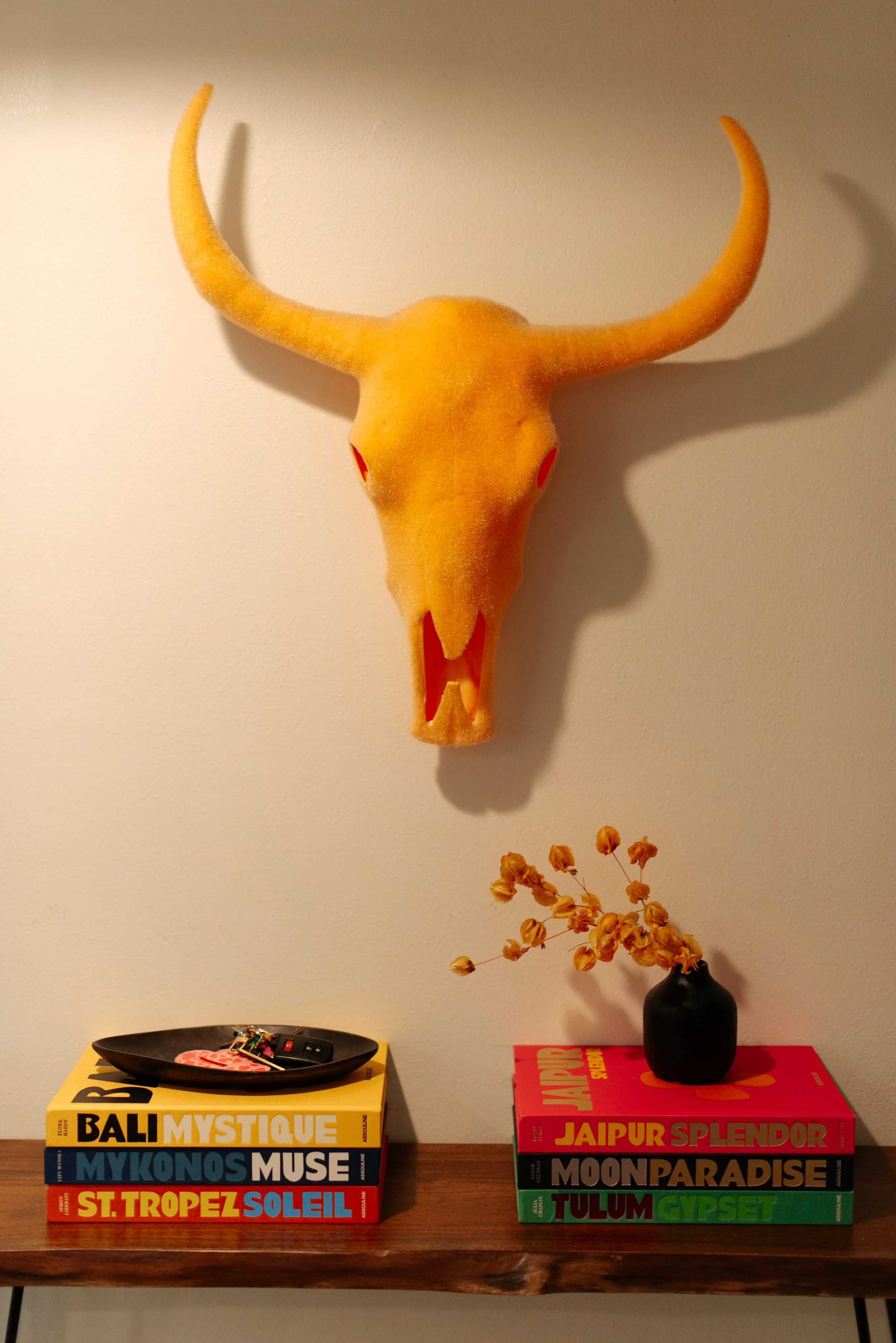 Decorative wall display with a large yellow object resembling a bull's skull with horns, positioned above two stacks of books on a wooden surface. The left stack includes travel books titled "BALI MYSTIQUE," "MYKONOS MUSE," and "ST. TROPEZ SOLEIL." T