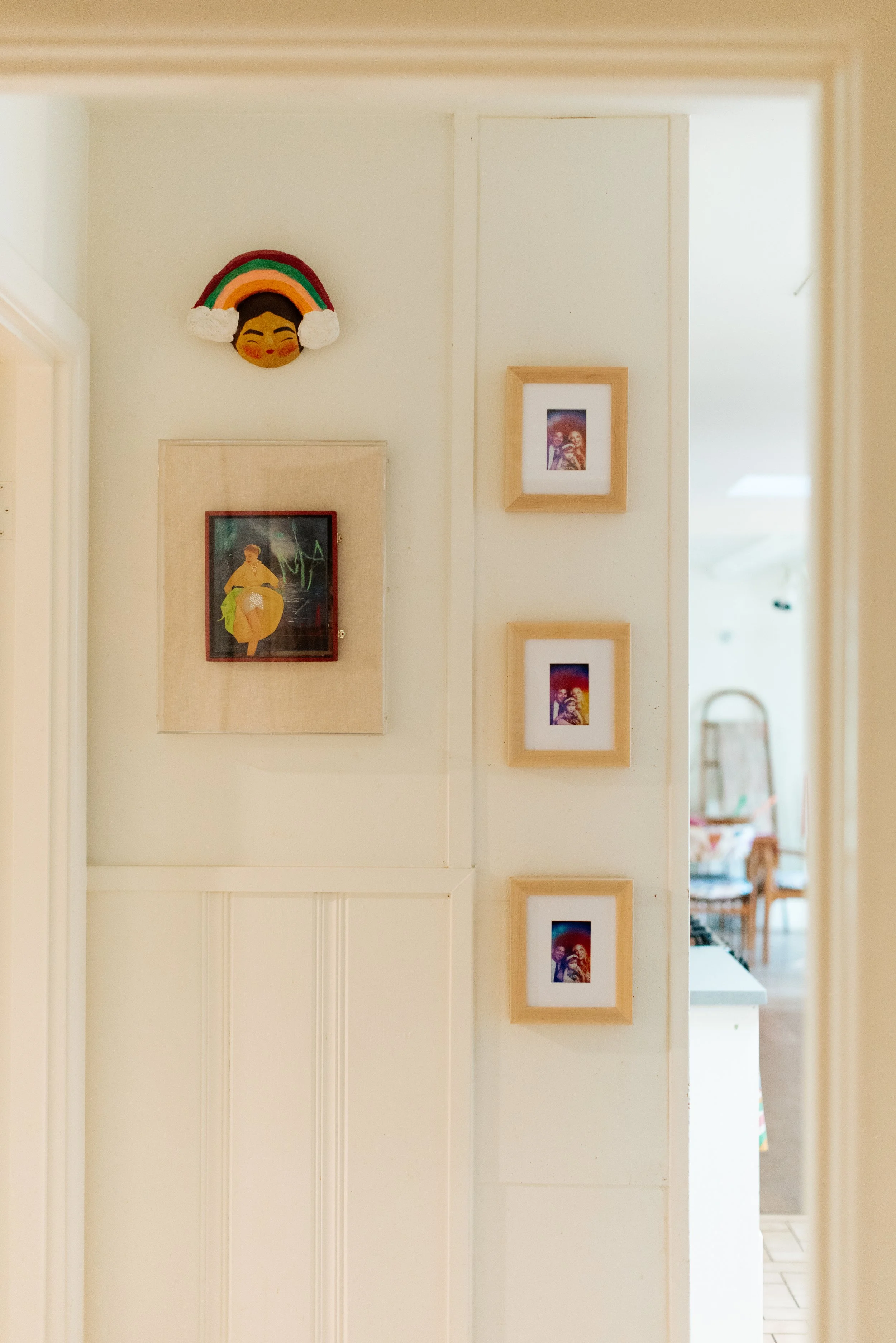Decorative wall with four framed photos, a colorful fabric face mask with a rainbow and cloud design, and a doorway leading to a kitchen or dining area.
