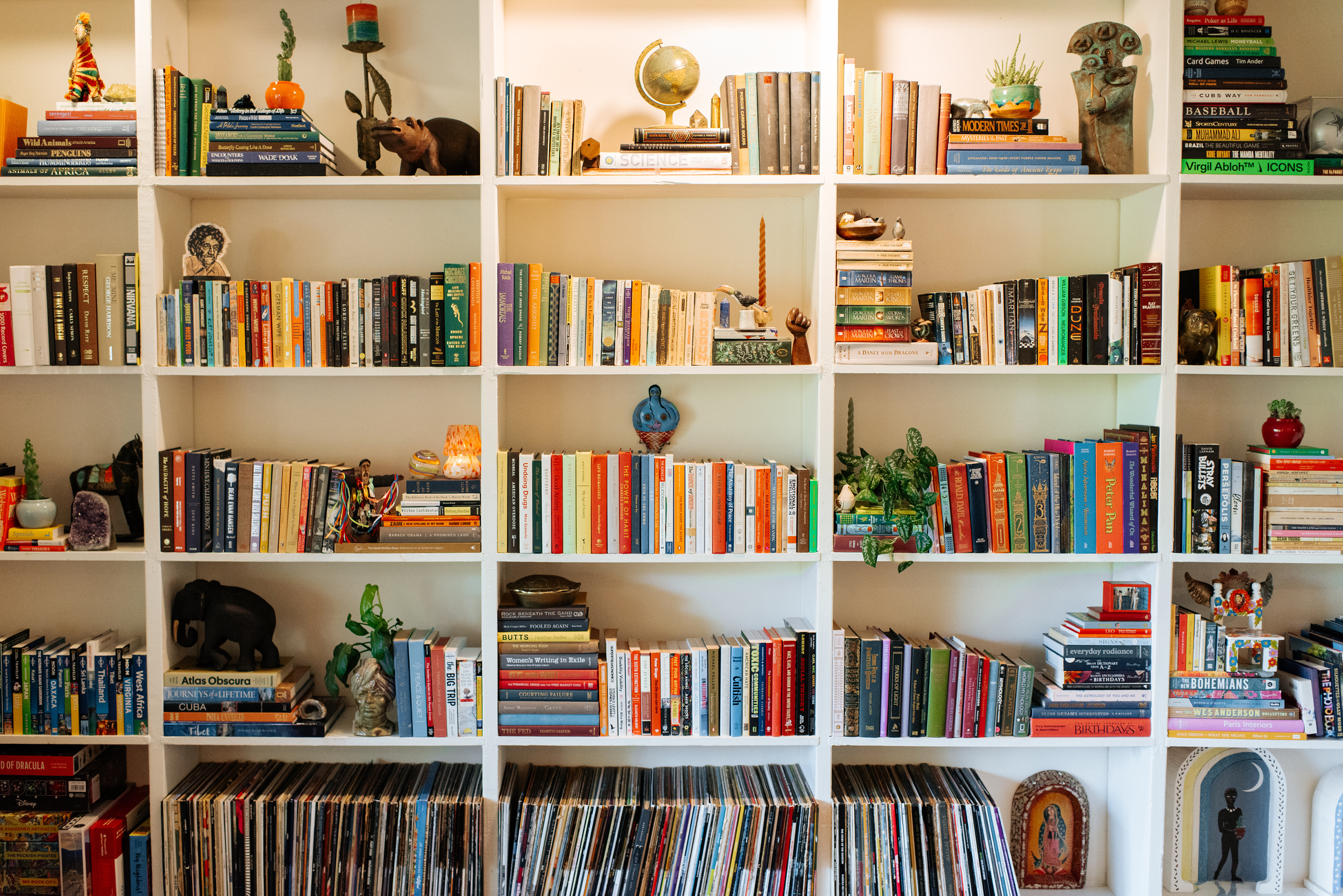 A white bookshelf filled with an array of colorful books and decorative objects, including plants, sculptures, and framed pictures.