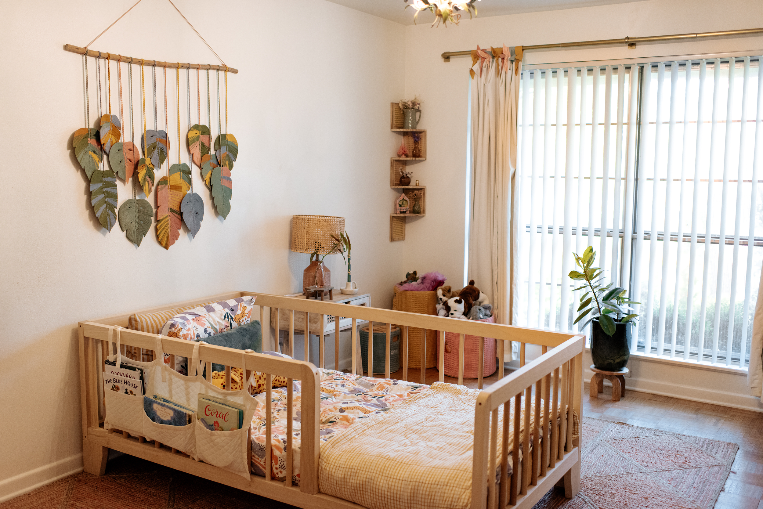 A cozy nursery with a wooden toddler bed, stuffed animals, and colorful wall decor, including a leaf-shaped wall hanging and shelves with decorative items, near a window with vertical blinds and curtains.