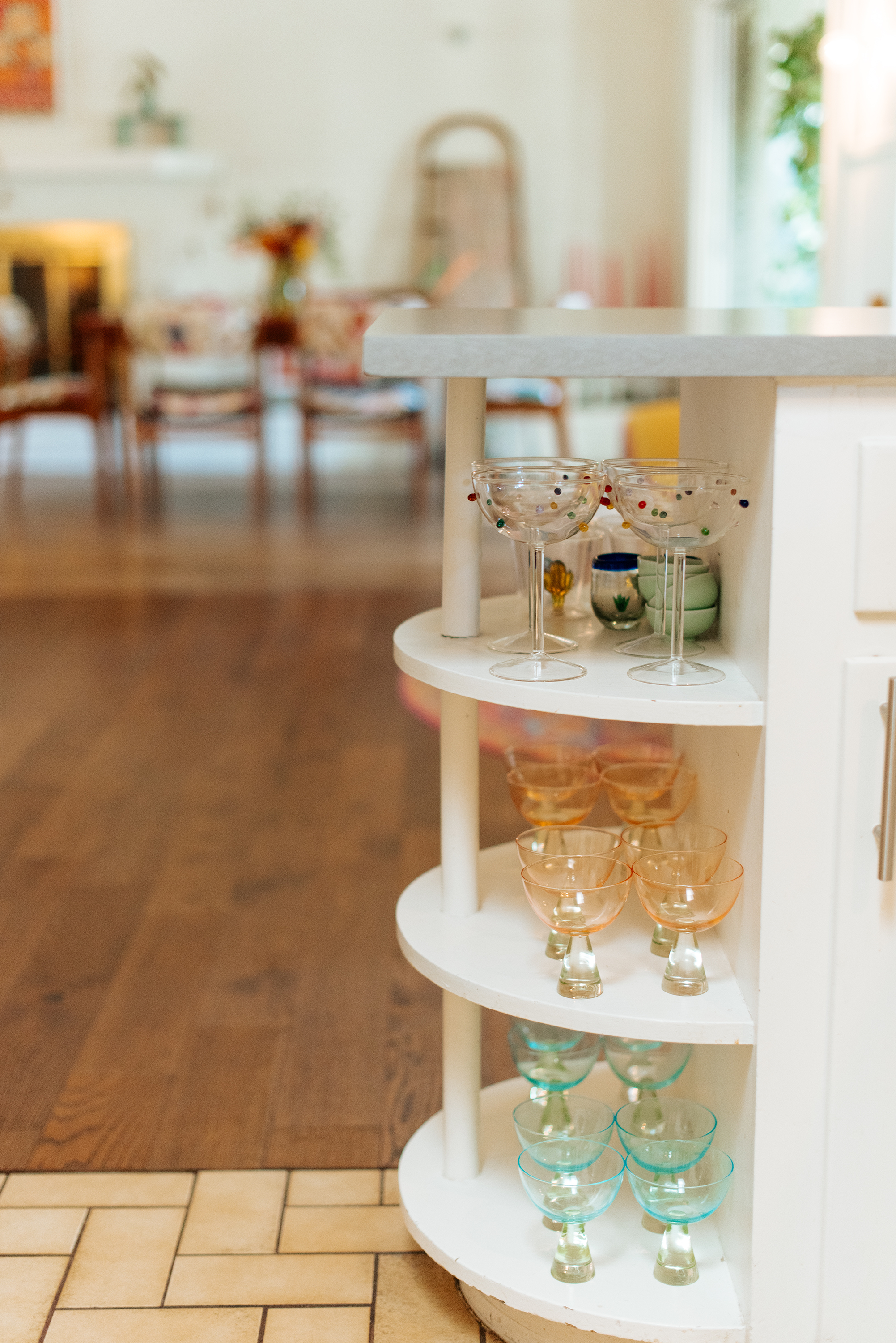 A white, curved, multi-tiered shelf displaying colorful glassware including martini glasses, cocktail glasses, and small bowls inside a bright room with wooden floors and blurred background furniture and decor.