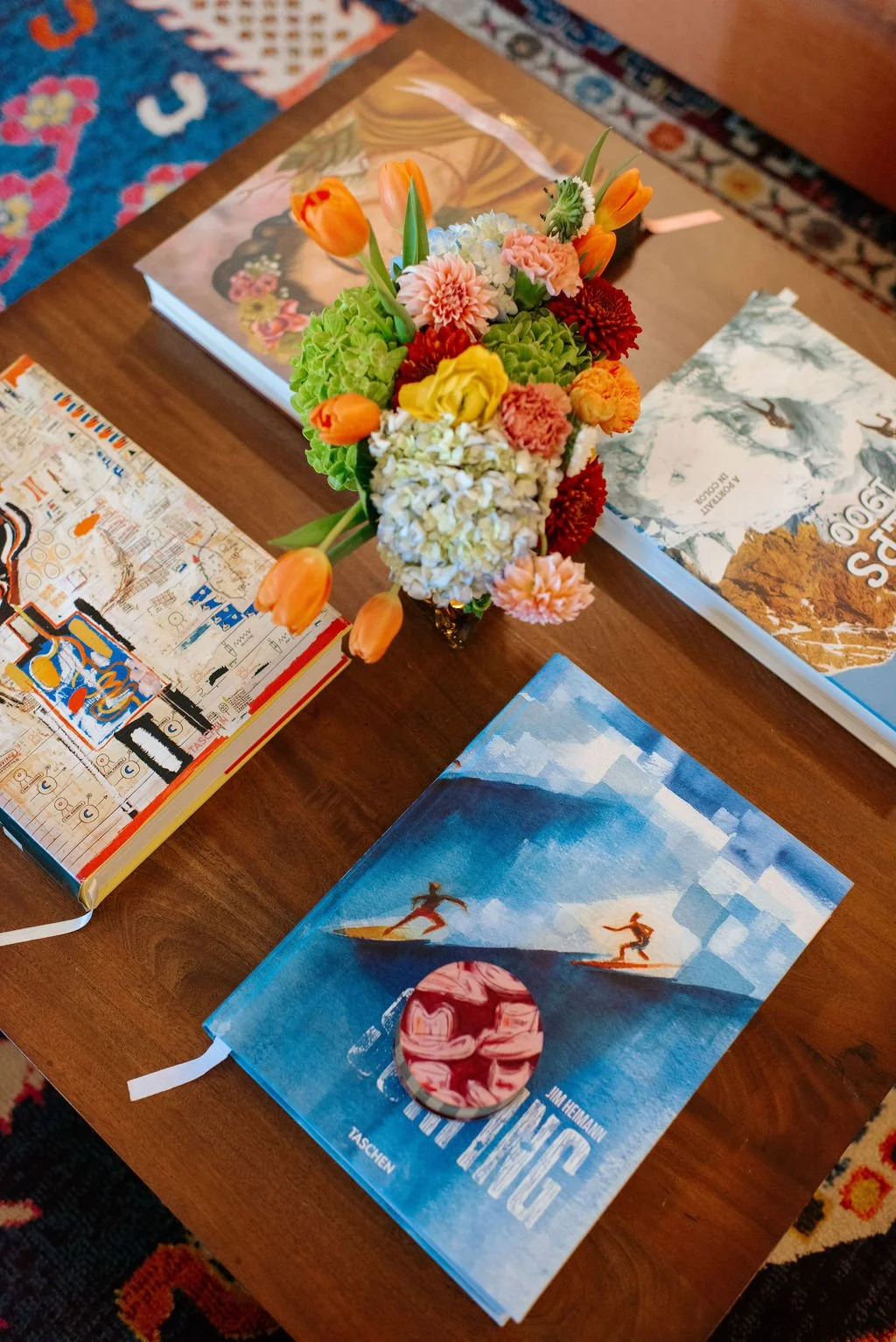 A wooden table with a colorful bouquet of flowers, three books, and a skincare or cosmetic compact. The bouquet includes orange tulips, pink and white carnations, and green hydrangeas. The books appear to be art or nature-themed, and the compact is p