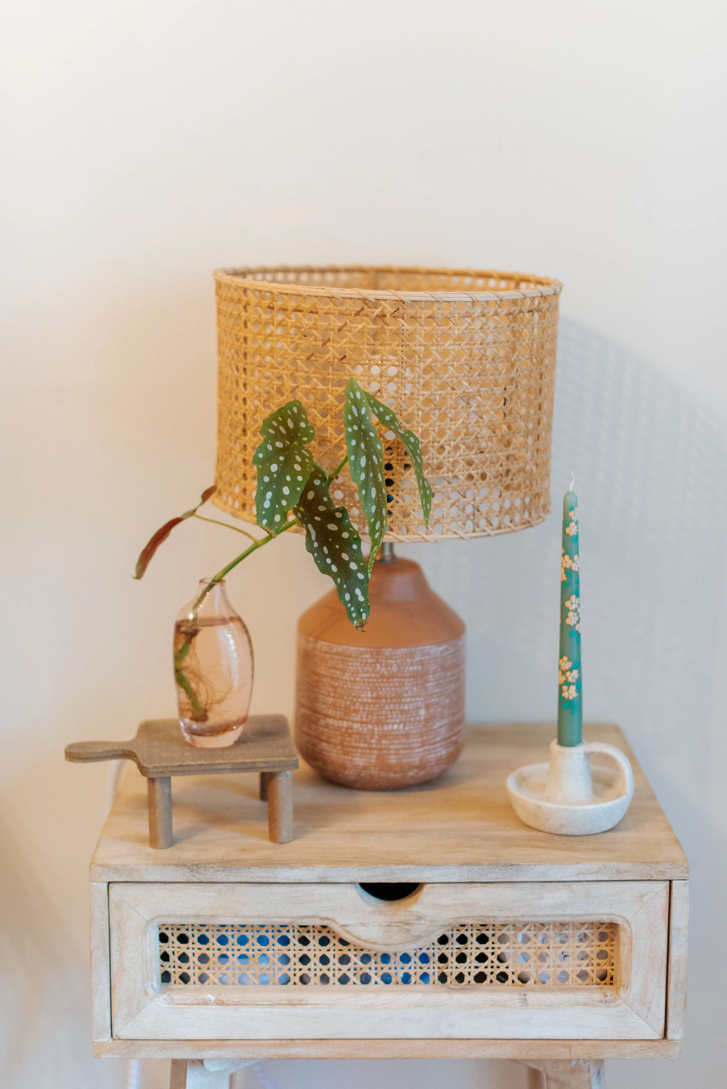 Decorative wooden side table with a small glass vase holding a leafy stem, a terracotta table lamp with a woven rattan shade, and a ceramic candlestick holder with a decorated candle on a light-colored wall background.