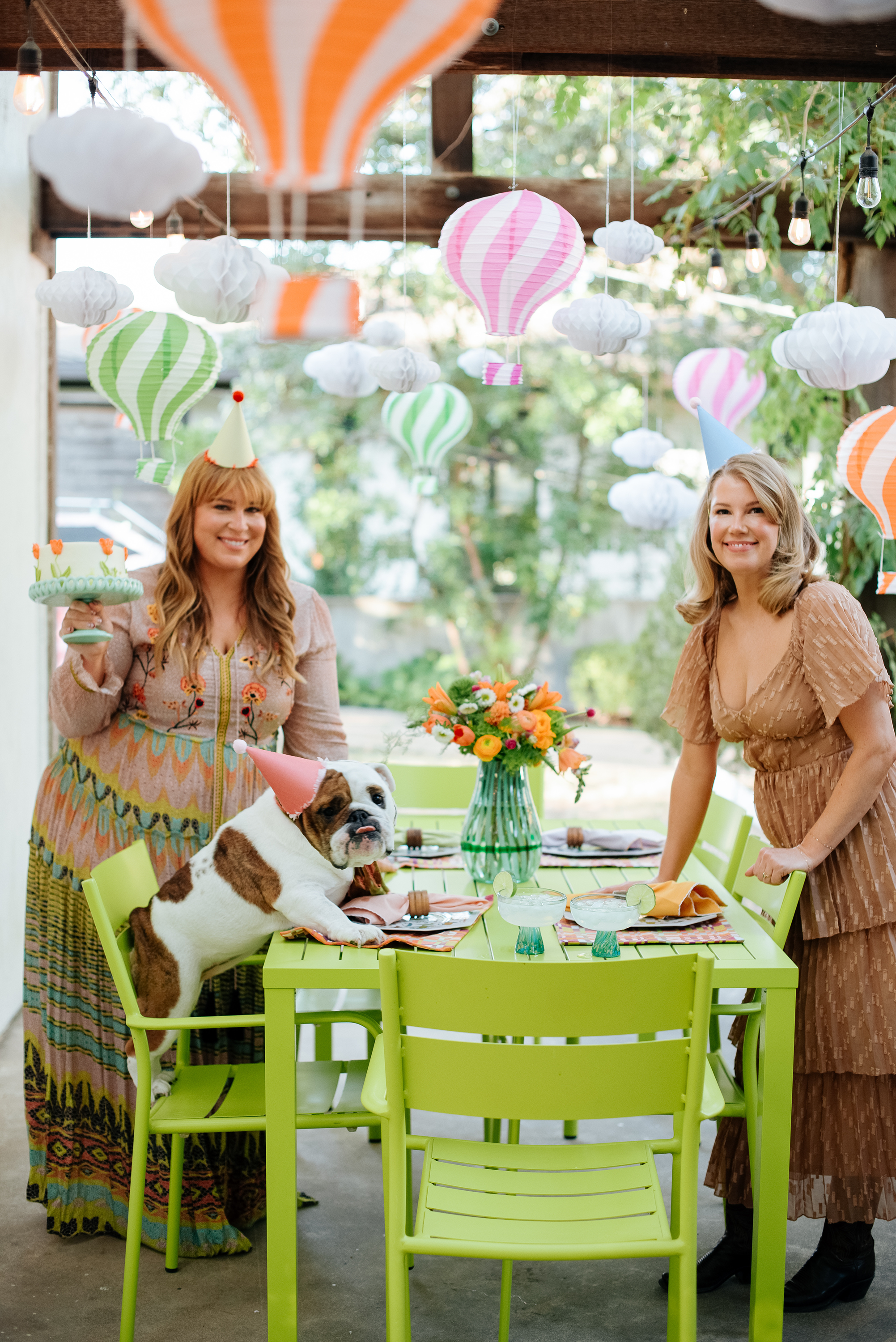 Two women celebrating a birthday with party hats, a dog in a party hat, colorful decorations, and a table set with drinks and flowers.