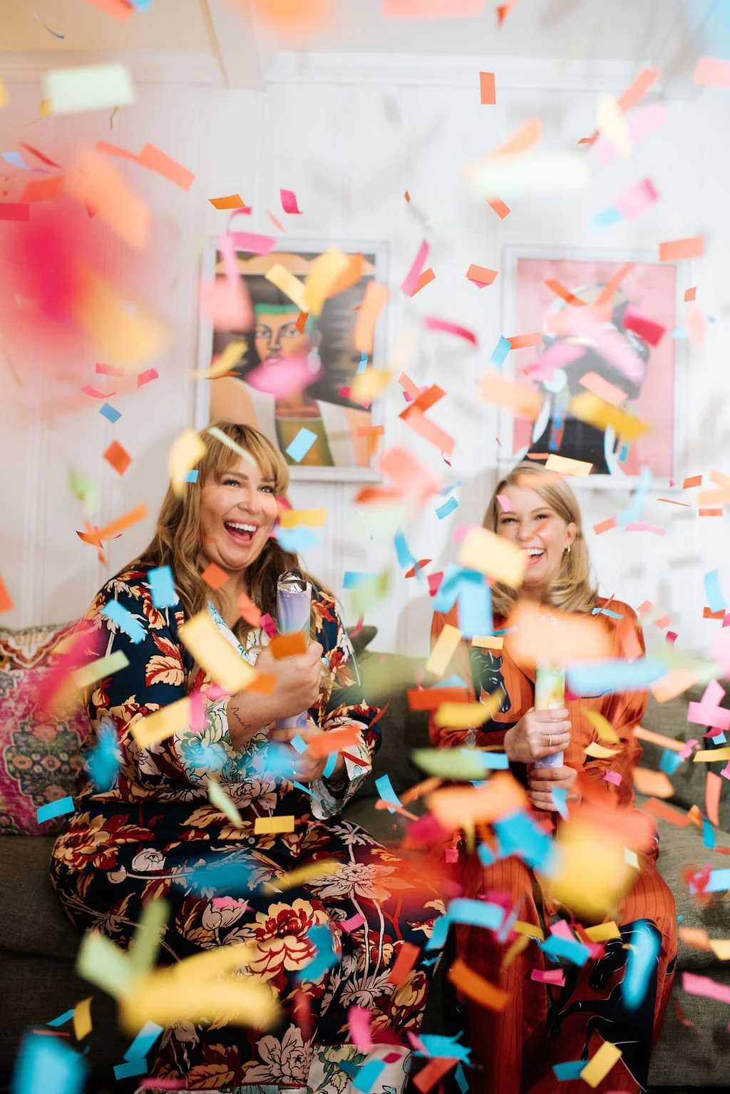 Two women celebrating indoors with colorful confetti falling around them, holding party poppers and smiling.