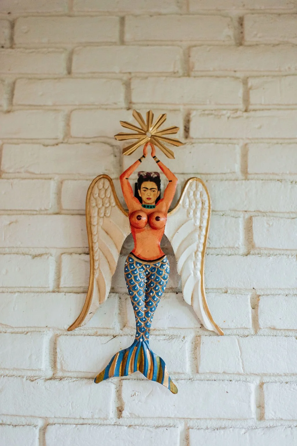 Decorative art piece of a woman with mermaid tail and angel wings, holding a starburst shape above her head, mounted on a white brick wall.