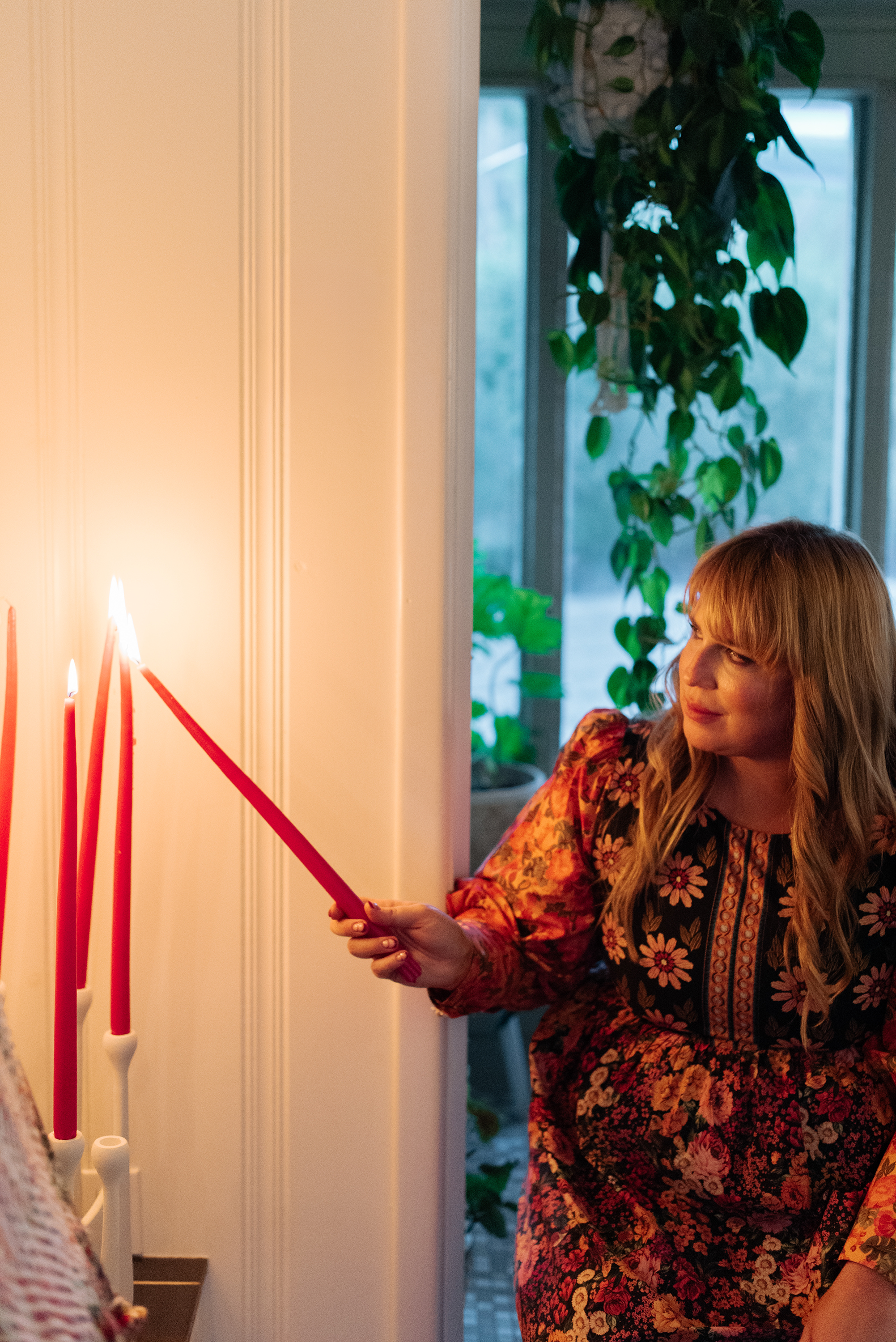 A woman lighting red candles on a wall-mounted candle holder in a cozy, indoor setting near a window with green plants in the background.